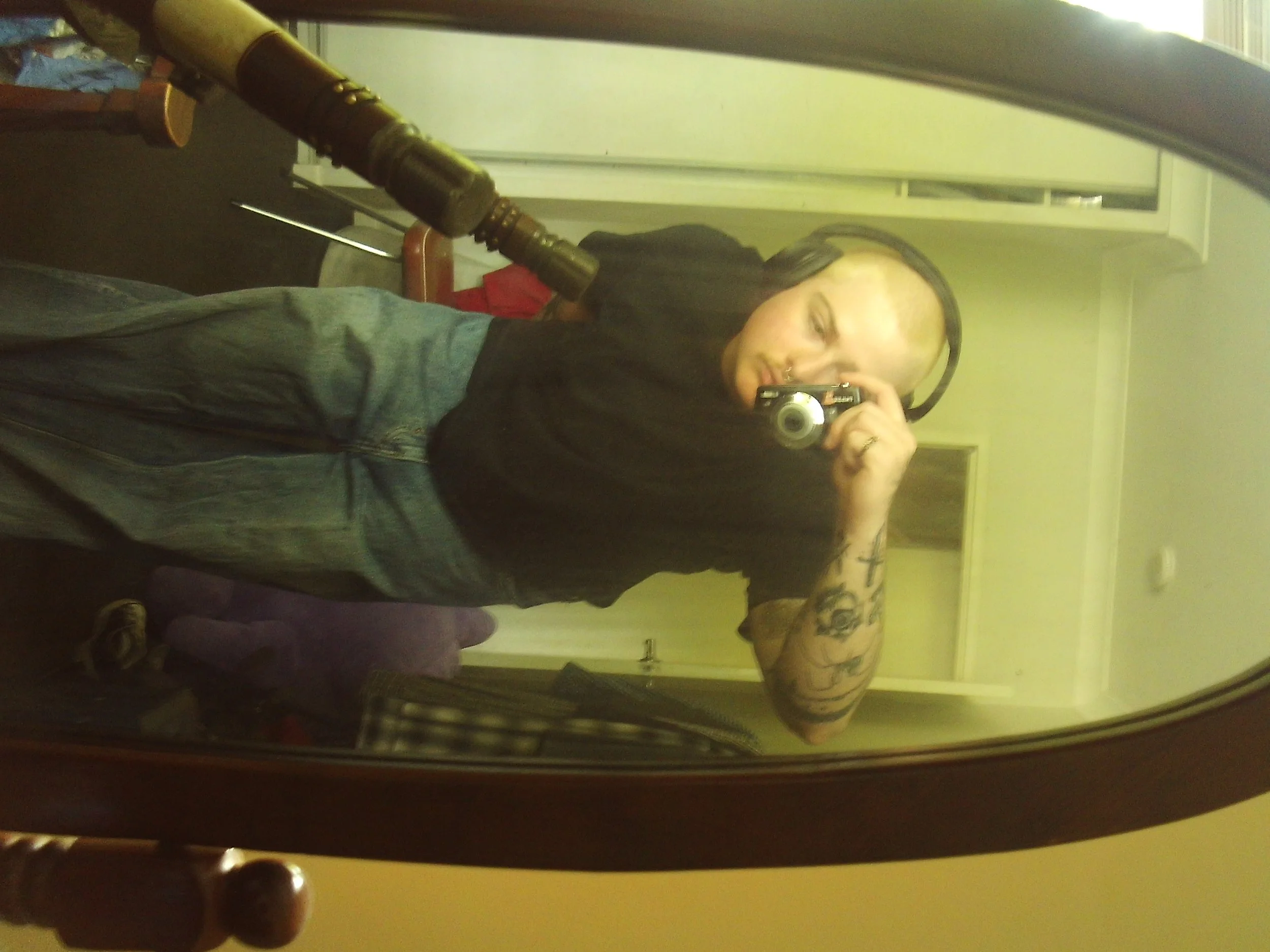 A woman with long dark hair, tattoos on her arm, and headphones around her neck taking a selfie in front of a large mirror. She is holding a camera and standing in a room with a yellow wall and a closet in the background.