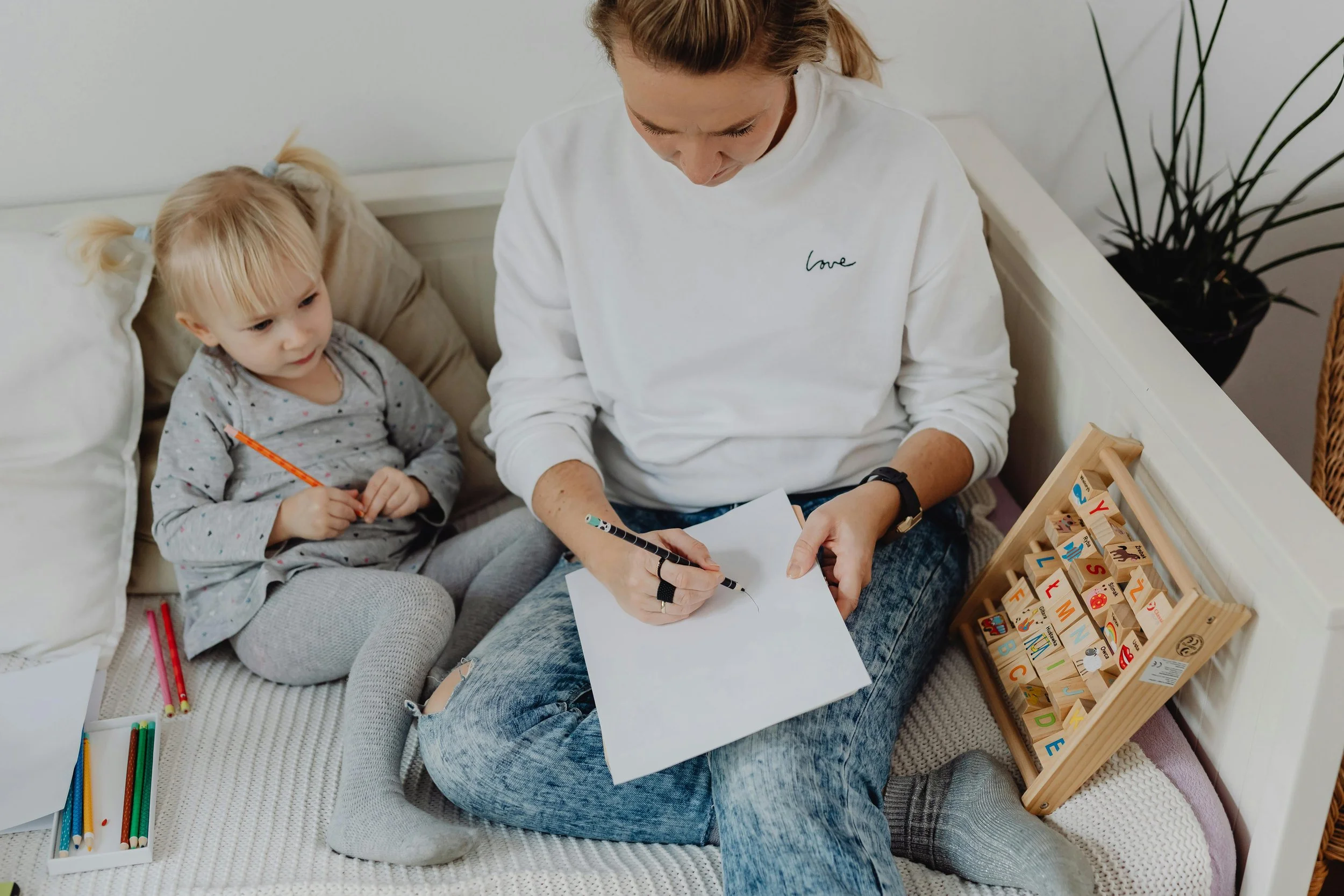 A young girl and an adult woman sitting on a bed with coloring supplies, including colored pencils and alphabet blocks, in a cozy room with a plant and a wooden shelf nearby.