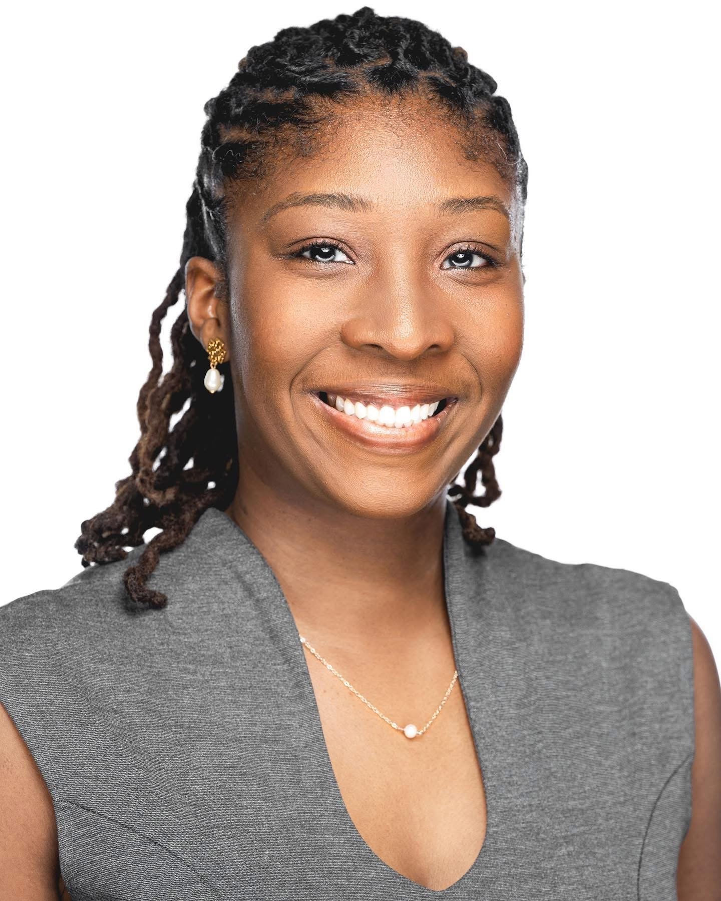 Close-up of a smiling woman with earrings, wearing a gray professional blazer and a pearl necklace, against a plain white background.