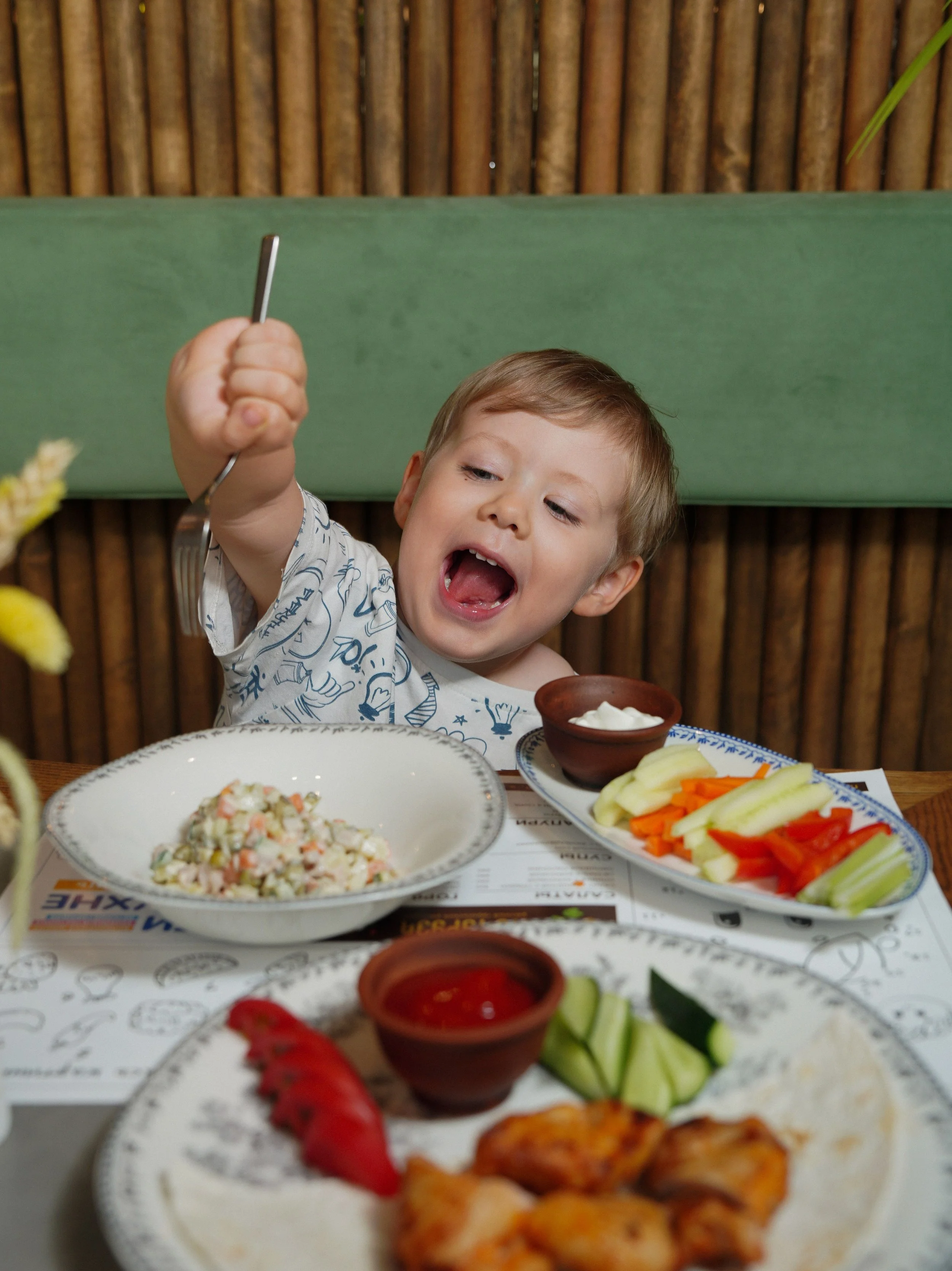 Young boy at a restaurant table with a variety of dishes, including salad, vegetables, and grilled chicken, holding a fork with a lively expression.