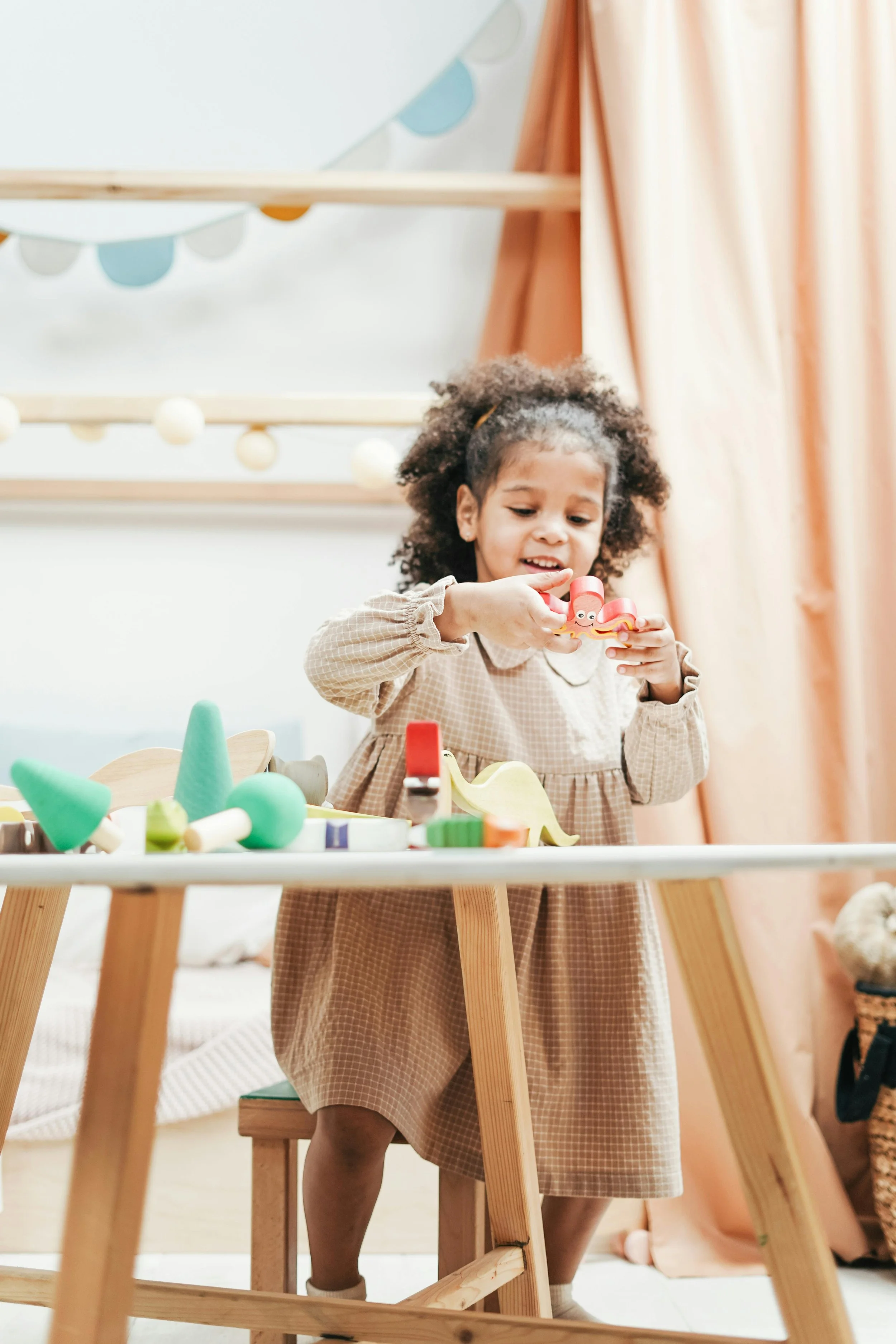 Young girl with curly hair and light brown skin playing with colorful wooden toys at a table in a cozy room with peach curtains.