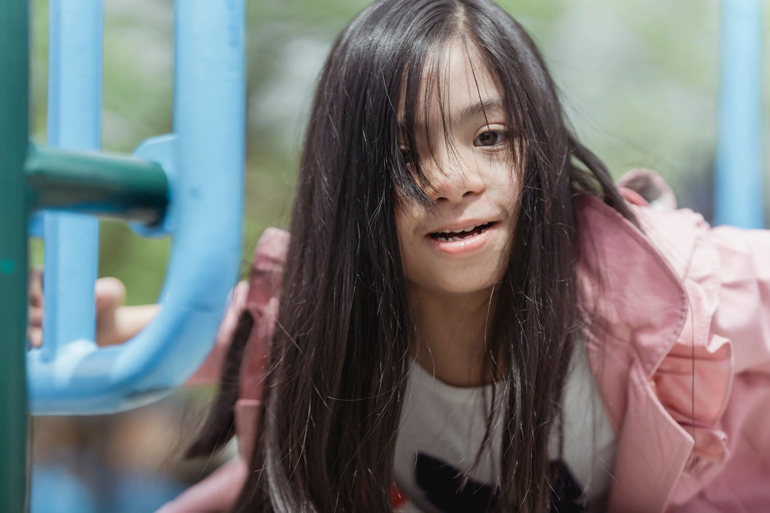 A young woman with long dark hair is leaning close to a blue playground structure, with her face partially obscured by her hair. She is smiling slightly outdoors.