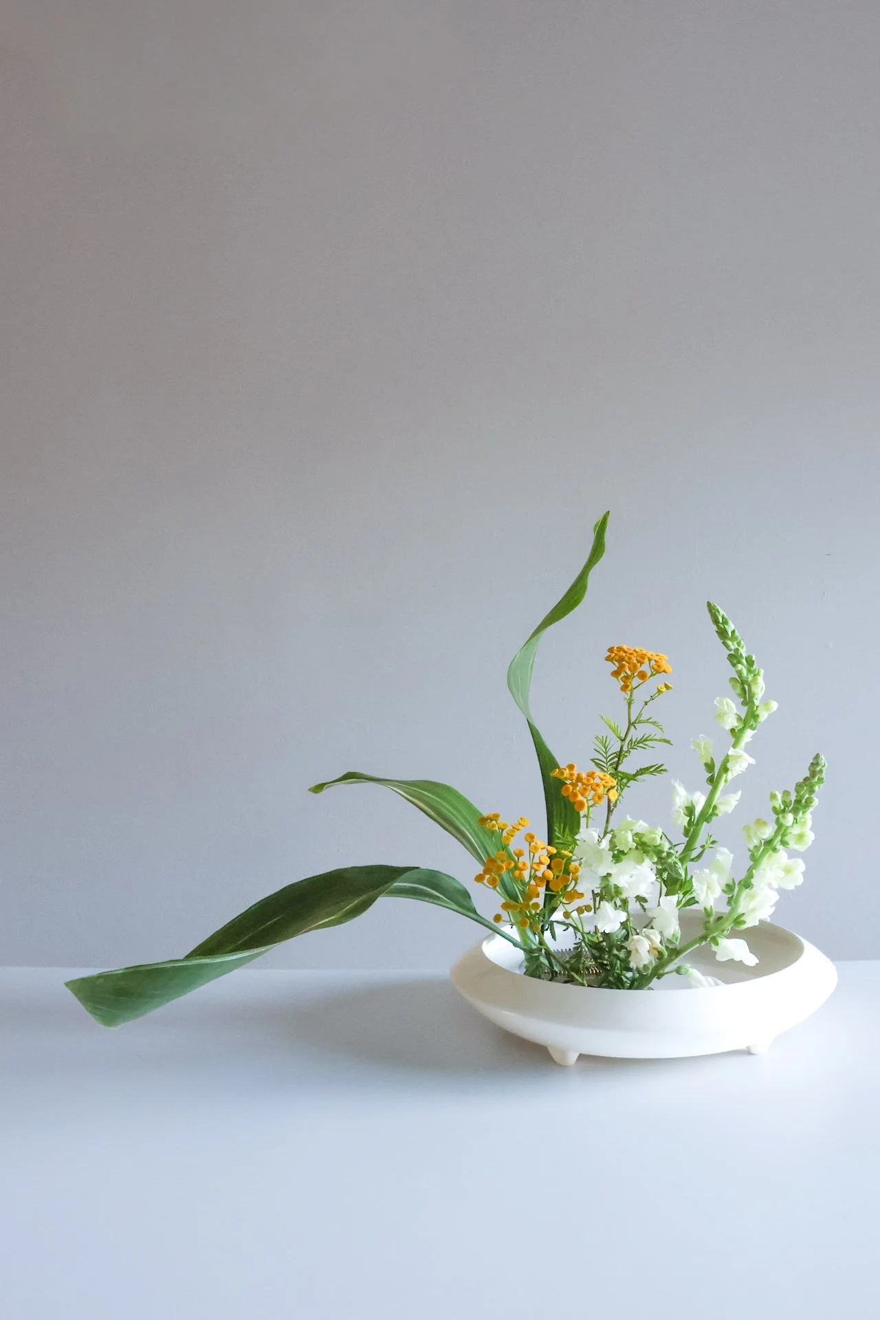 A white ceramic vase containing various flowers and greenery, placed on a white surface against a gray background.