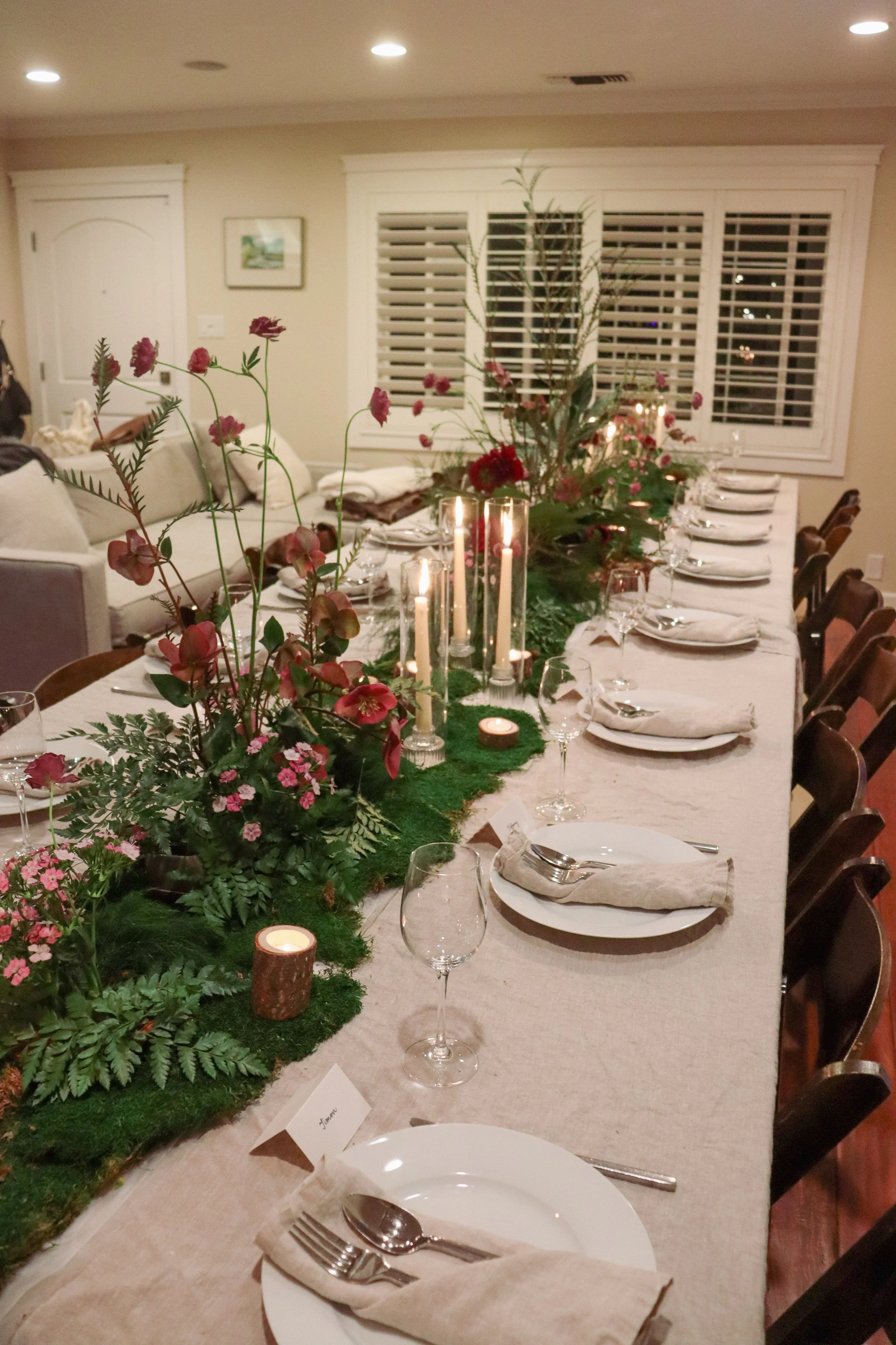 A long dining table decorated with floral arrangements, greenery, and lit candles, set for a dinner gathering in a room with beige walls, white window shutters, and table settings including plates, glasses, and silverware.