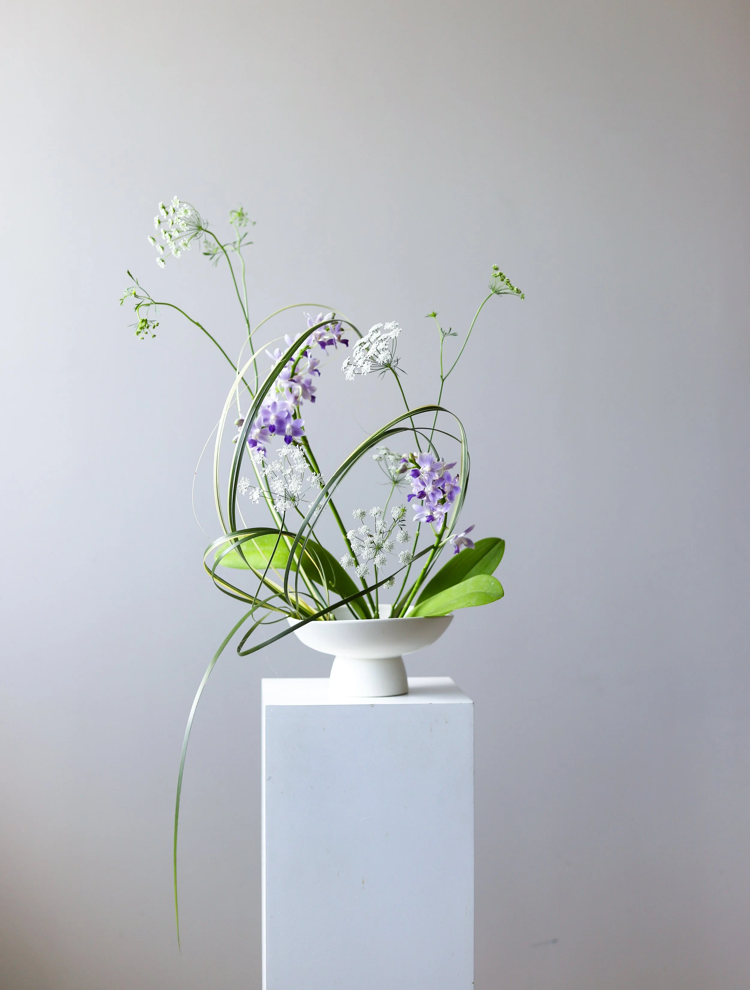 A white vase with green and purple flowers and greenery, displayed on a white pedestal against a plain light gray background.