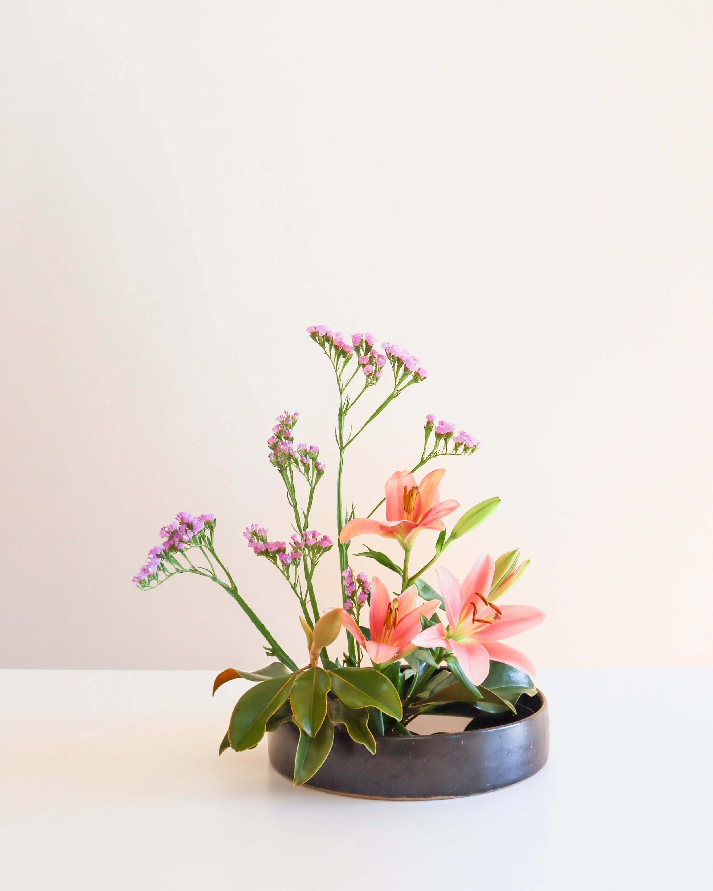 Flower arrangement with pink lilies, purple statice, and green foliage in a black bowl, set against a light background.