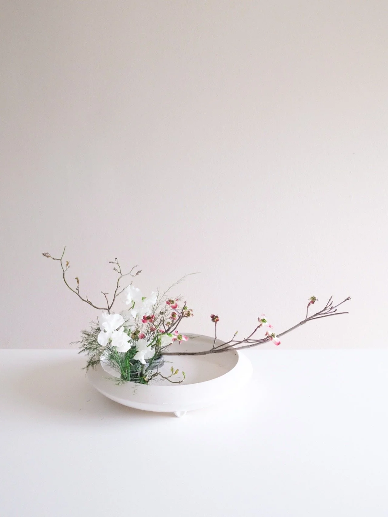 A minimalist flower arrangement in white ceramic vase with white and pink flowers and bare branches on a white surface against a cream wall.