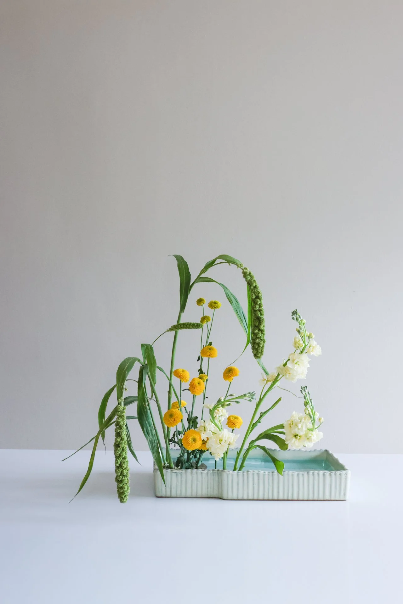 A rectangular ceramic vase with white and turquoise glaze holds a mixed arrangement of yellow, white, and green flowers and foliage, set against a plain light gray background.