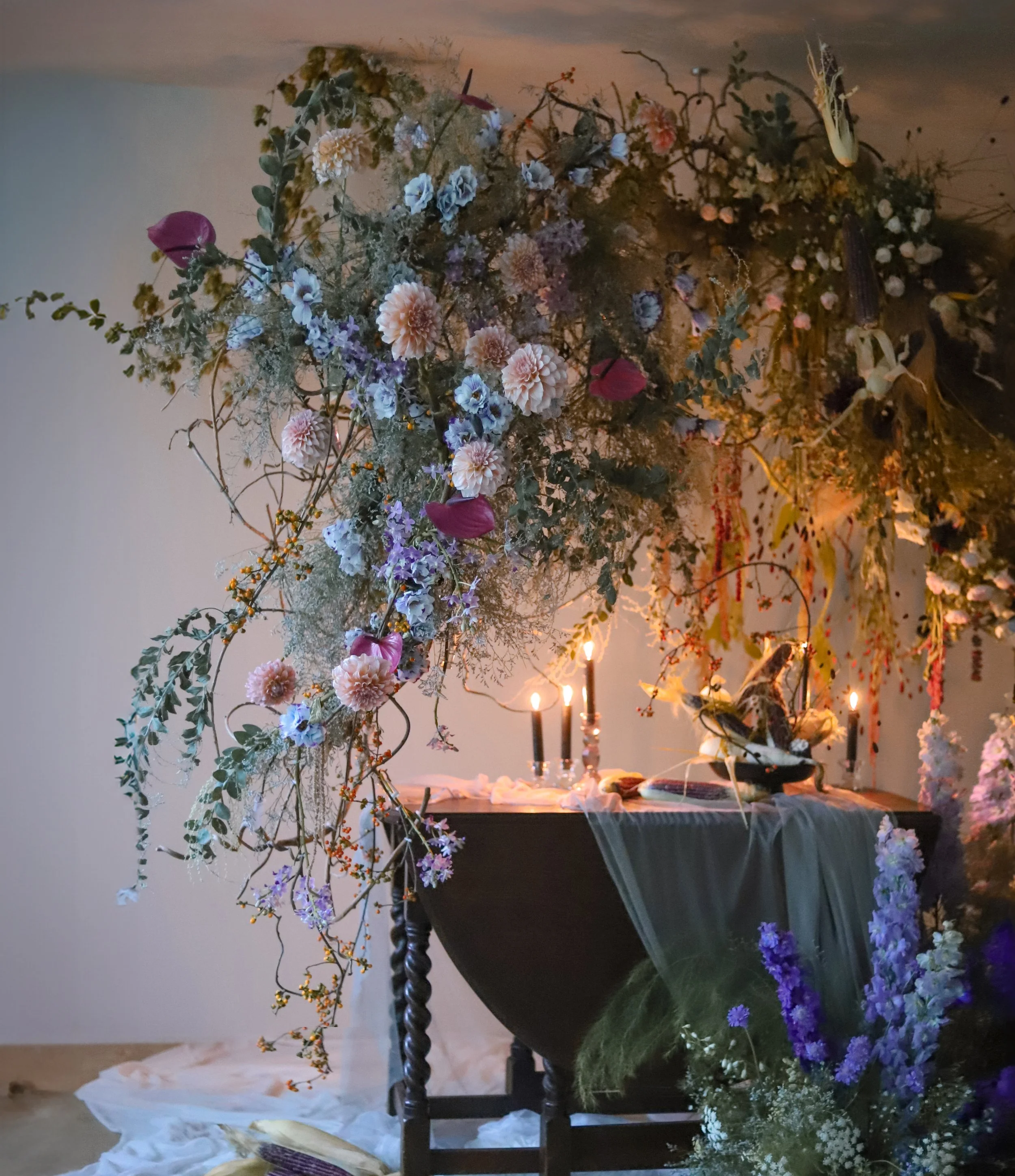 A dark-colored vase with a large, elaborate floral arrangement featuring pink dahlia flowers, purple and blue flowers, greenery, and branches with small berries. The arrangement is on a black table with a white cloth, lit by candlelight, with additional flowers and greenery at the base.