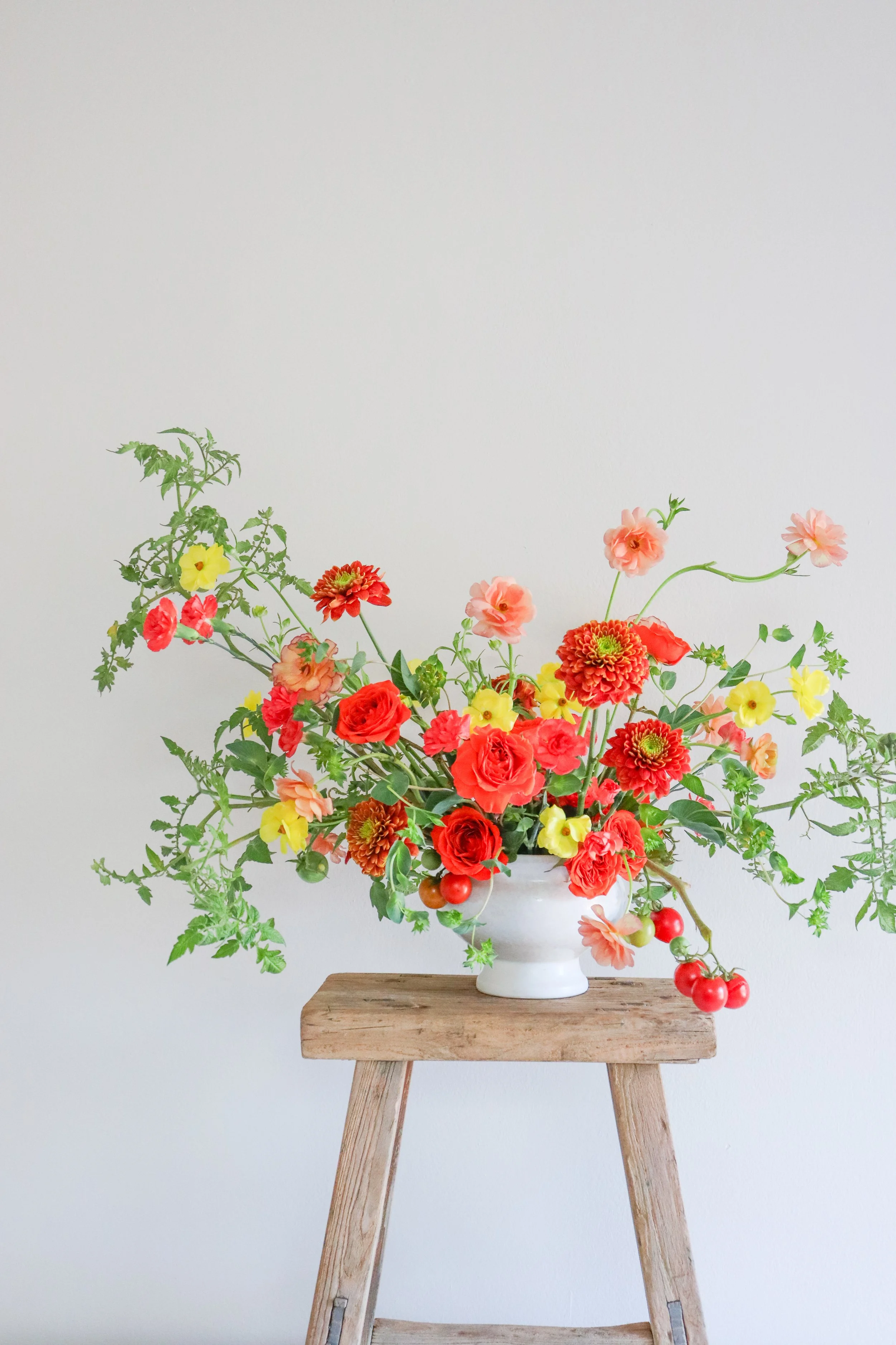 A floral arrangement in a white vase on a wooden stool against a plain light-colored wall. The arrangement includes red, pink, yellow flowers, and green foliage, with a chain of small red tomatoes cascading from the arrangement.