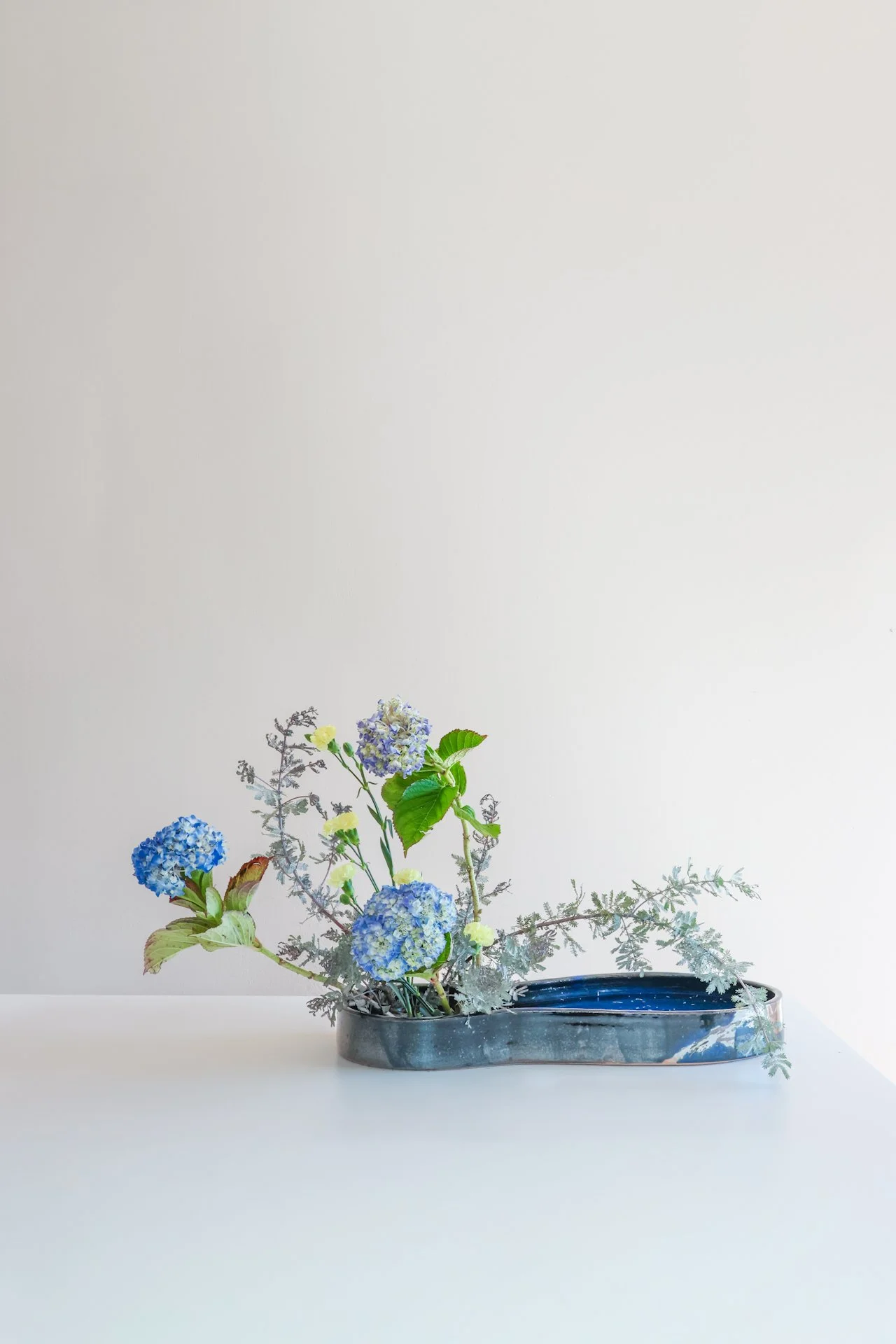 A floral arrangement in a long, oval-shaped ceramic vase placed on a white surface against a plain light-colored background. The arrangement includes blue hydrangeas, yellow flowers, green leaves, and various dried foliage.