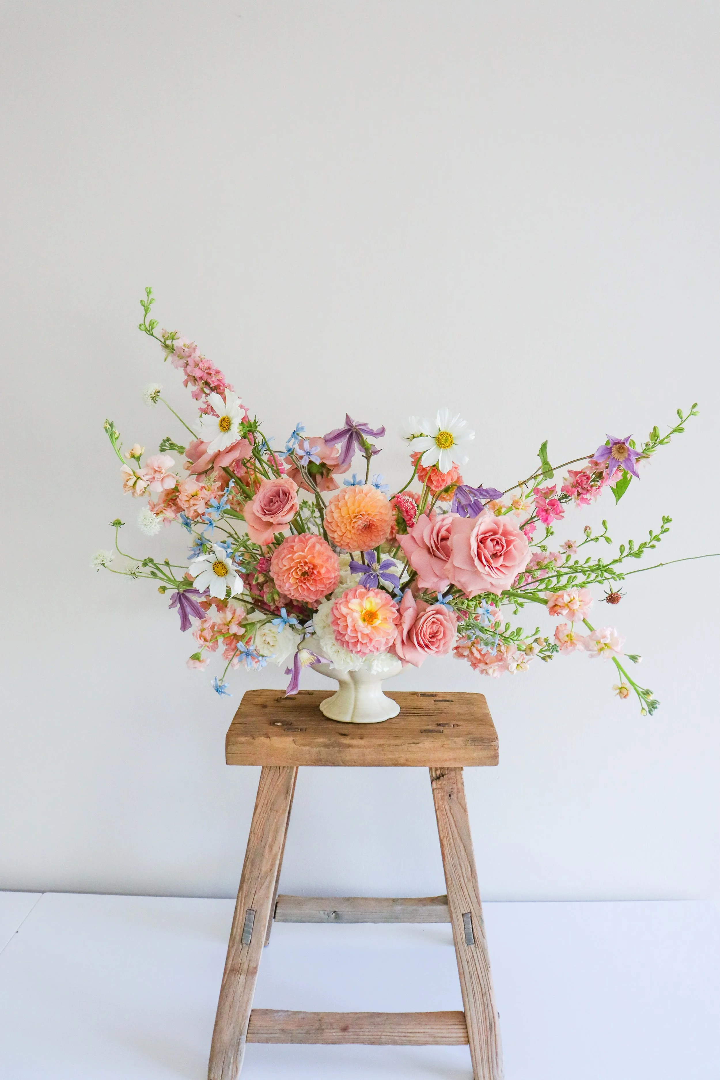 A large bouquet of pink, white, and purple flowers arranged in a beige vase on a wooden stool against a plain white wall background.