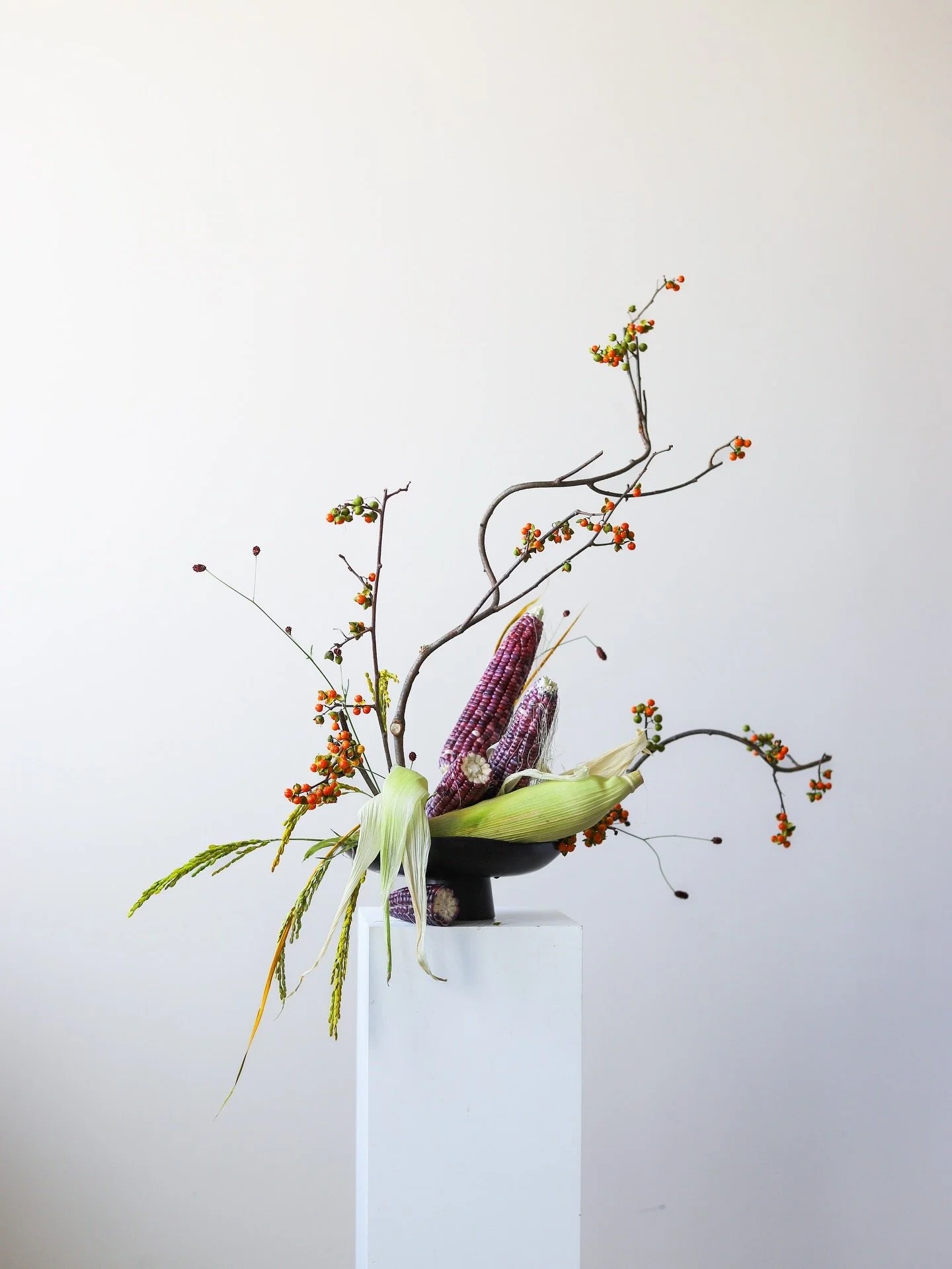 A flower arrangement featuring branches with small orange and green berries, purple and white corn cobs, yellow-green leaves, and long wheat stalks, displayed in a black vase on a white pedestal against a plain, light-colored background.