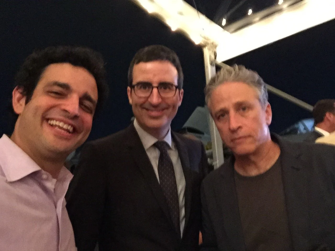 Three men taking a selfie at an event, with a dark background and stage lighting.