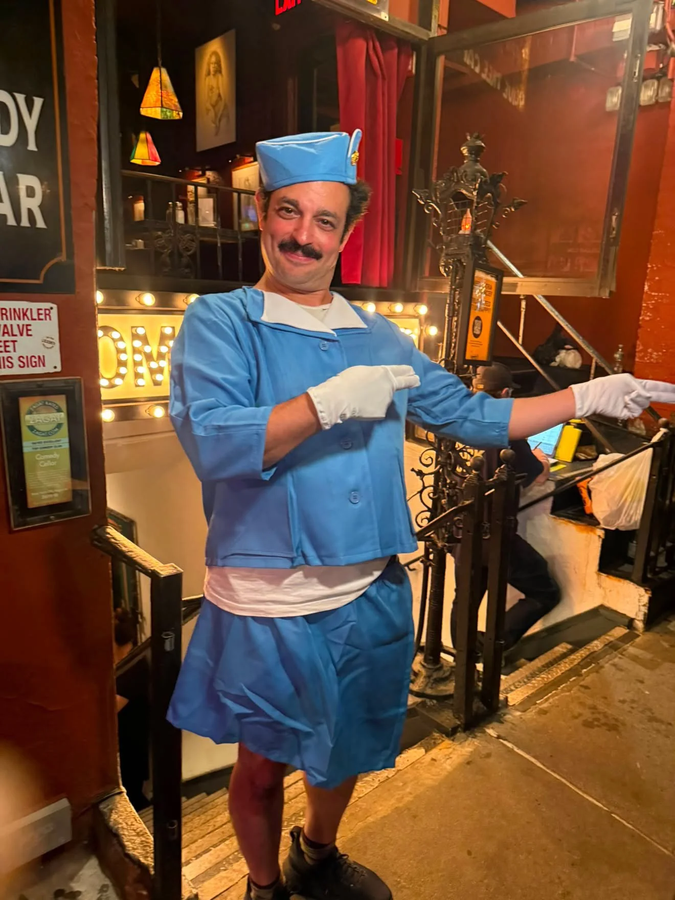 A man dressed in a blue Boston Cream Pie costume, complete with a pudding cup hat, striped socks, and black shoes, standing on outdoor stairs at night, smiling and pointing.