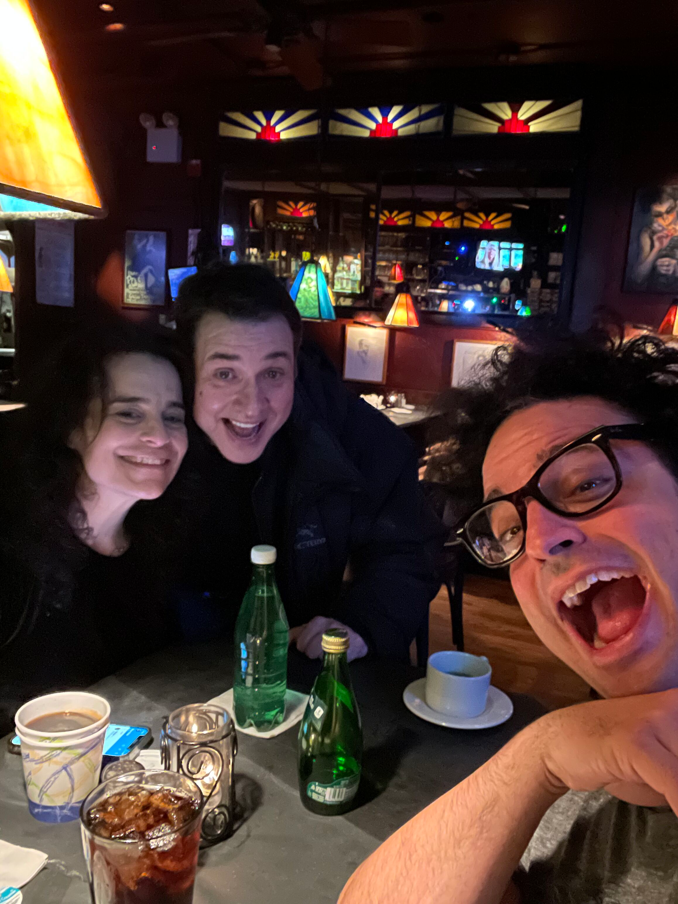 Four people are sitting around a table in a dimly lit restaurant or bar, smiling and enjoying drinks. The table has three bottles of sparkling water, two glasses of soda with ice, a cup of coffee, and a bowl of food. The background features decorativ