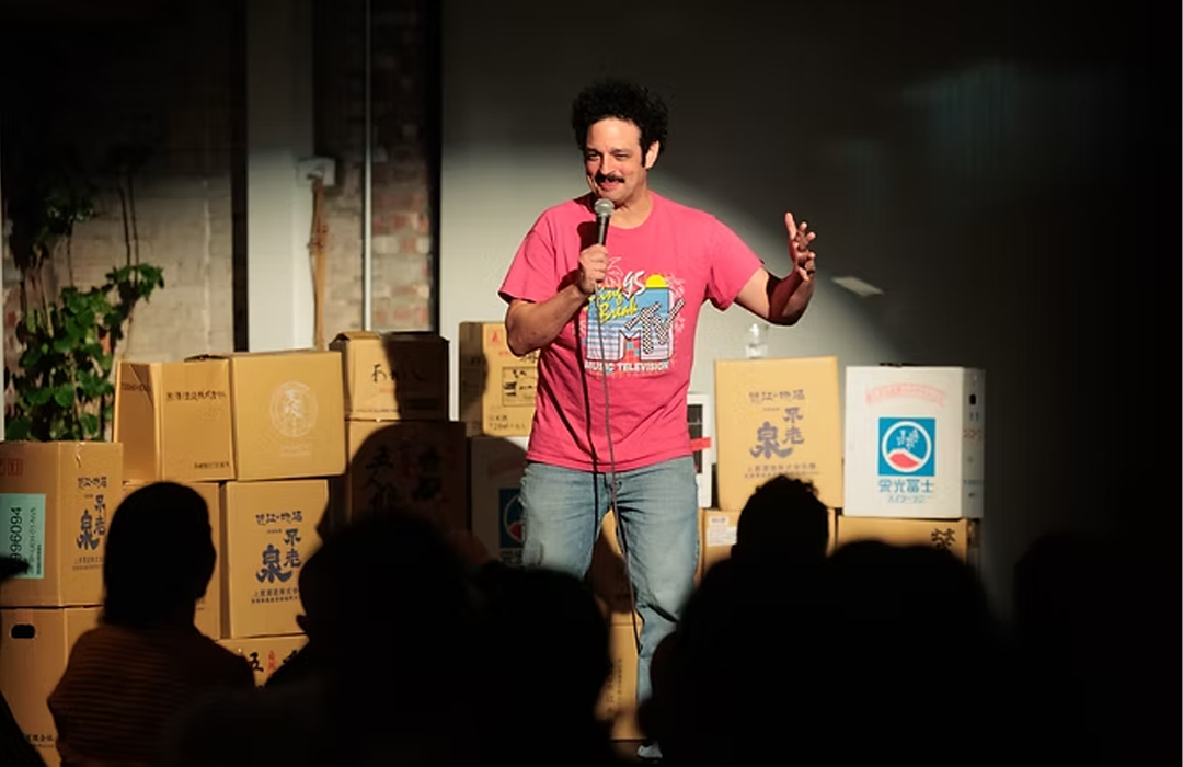 A man with dark curly hair and a mustache, wearing a pink T-shirt and blue jeans, standing on stage holding a microphone and speaking or performing in front of an audience. Behind him are boxes stacked in the background.