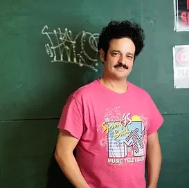 Man with curly hair and a mustache wearing a pink 'MTV Music Television' t-shirt, standing in front of a chalkboard with chalk writing.