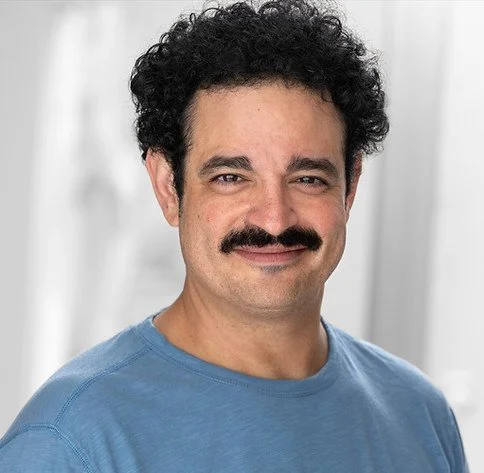 Headshot of a man with curly hair, a mustache, wearing a blue t-shirt, smiling, with a light-colored background.