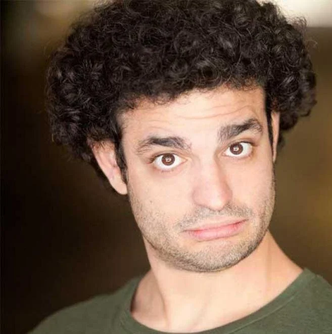 Close-up of a young man with curly dark hair, light skin, and wearing a green shirt, looking directly at the camera with a neutral expression.