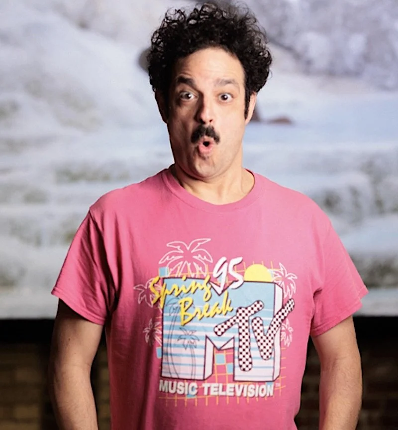 A man with curly dark hair and a mustache wearing a pink T-shirt with a tropical beach design, the words 'Sun's Beach,' and 'Music Television' on it.