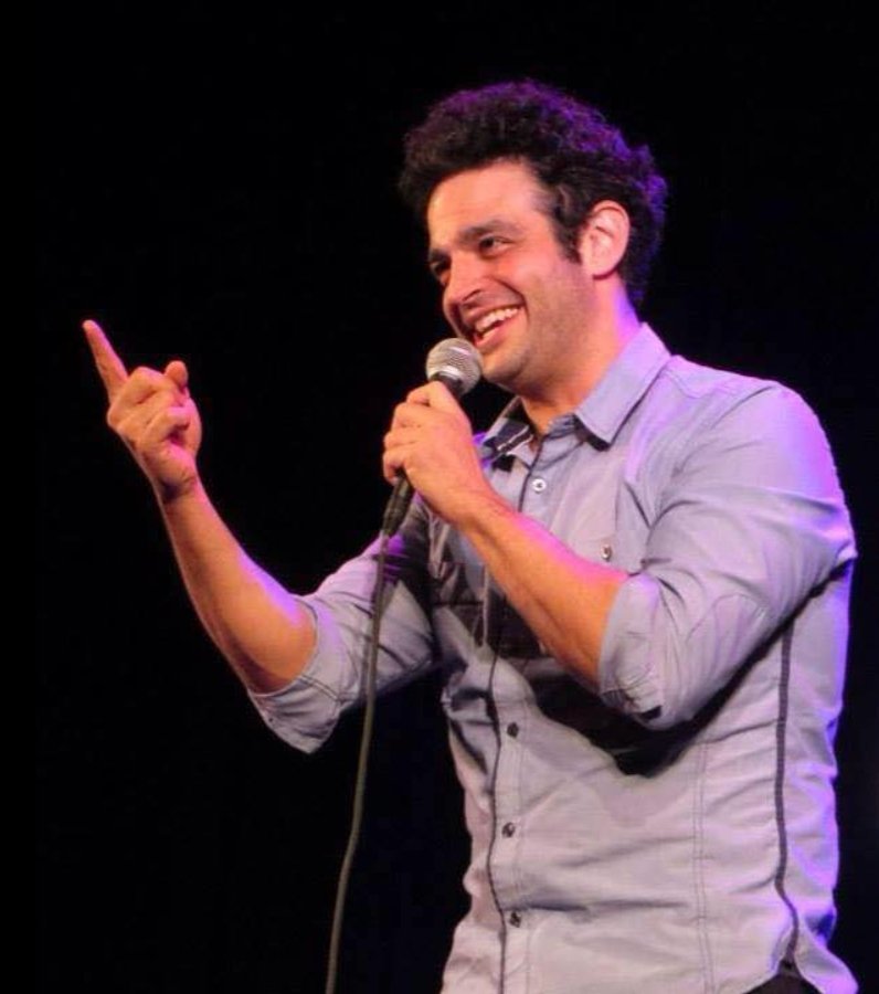 Man performing stand-up comedy on stage, holding a microphone and smiling.
