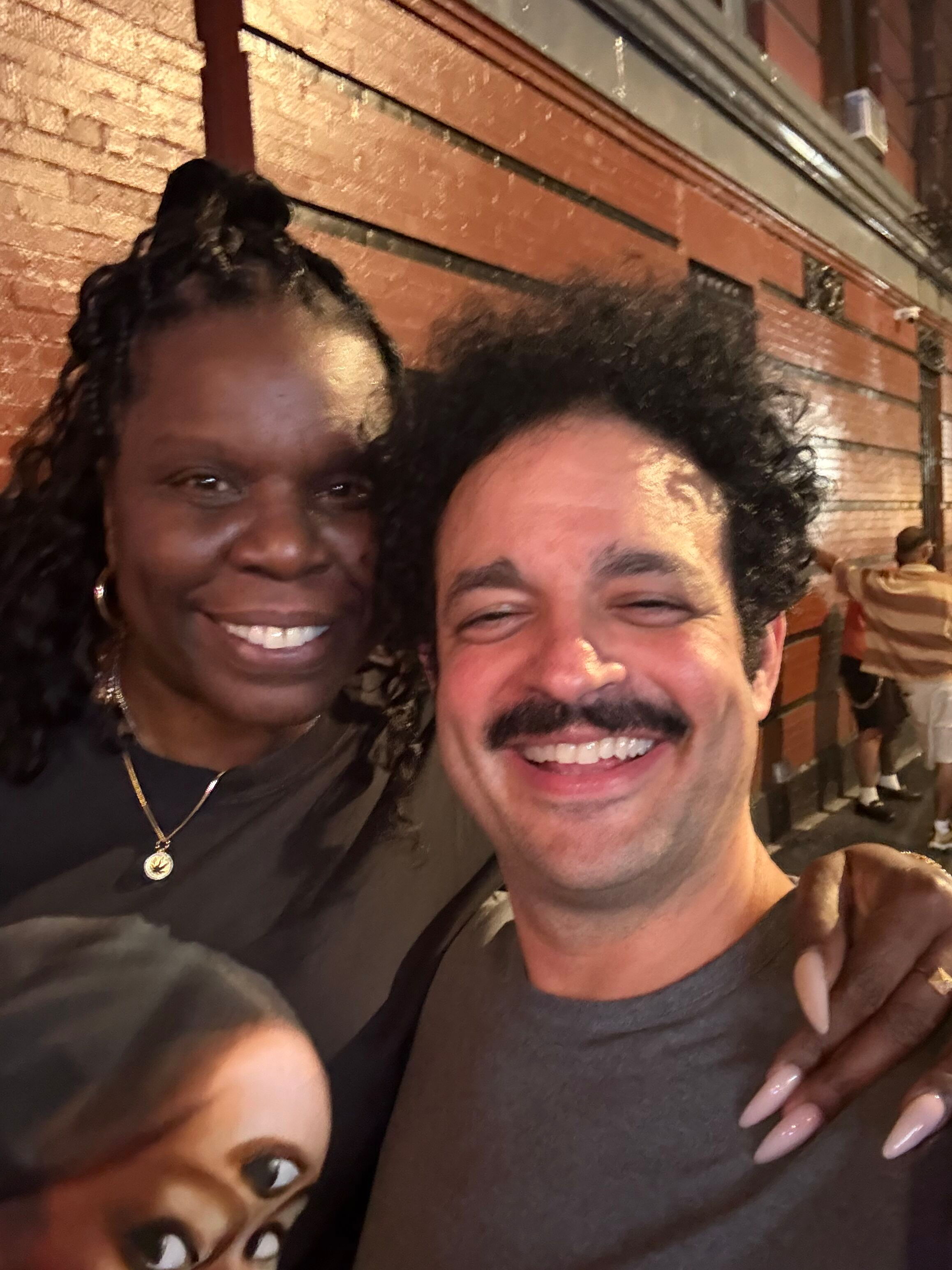 Close-up of smiling woman with dark braided hair and earrings, and smiling man with dark curly hair, in front of a brick wall at night.