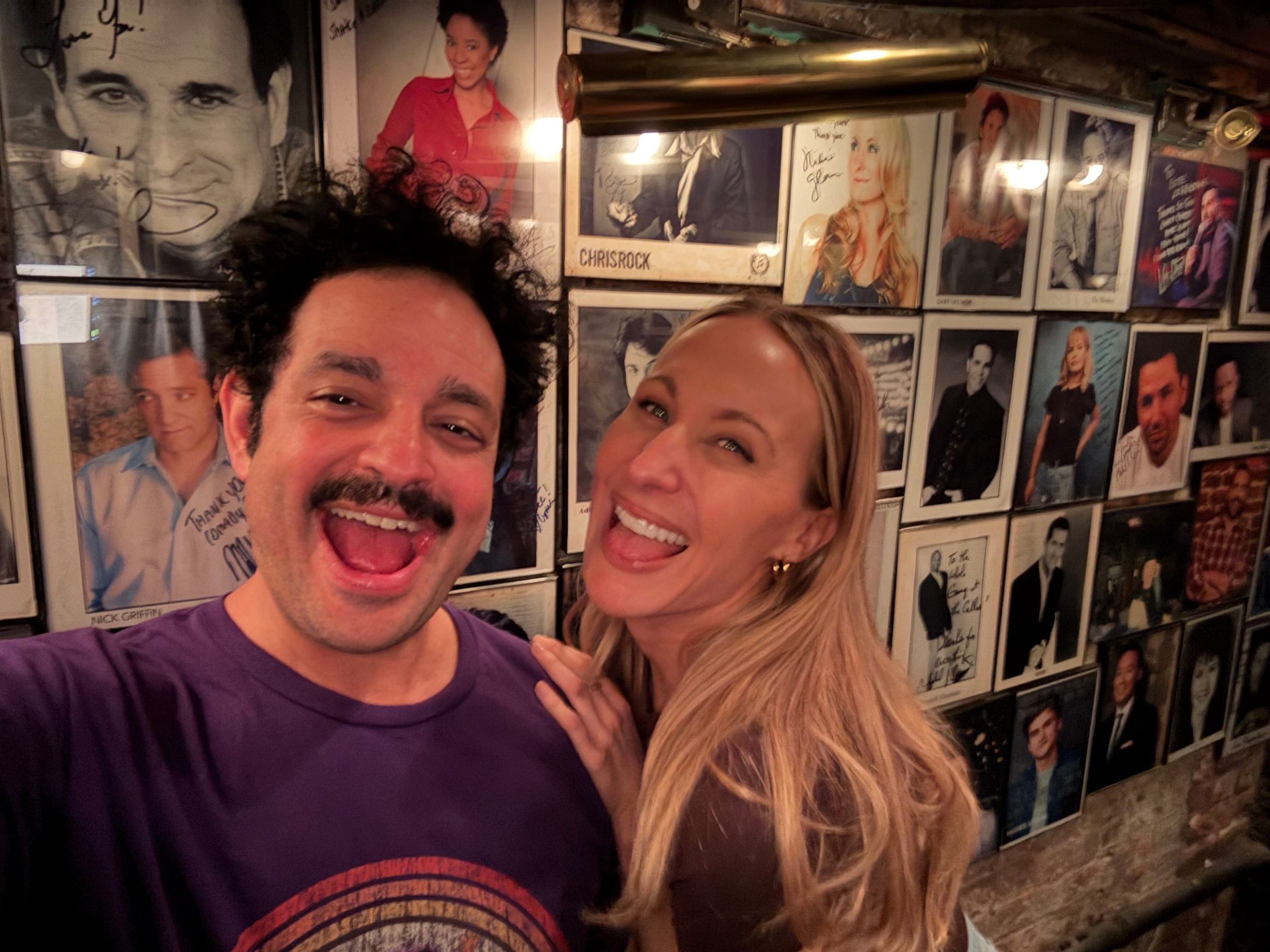 A smiling man with dark curly hair and a beard, and a smiling woman with long blonde hair, pose for a selfie in front of a wall decorated with framed photographs of celebrities and signed memorabilia.