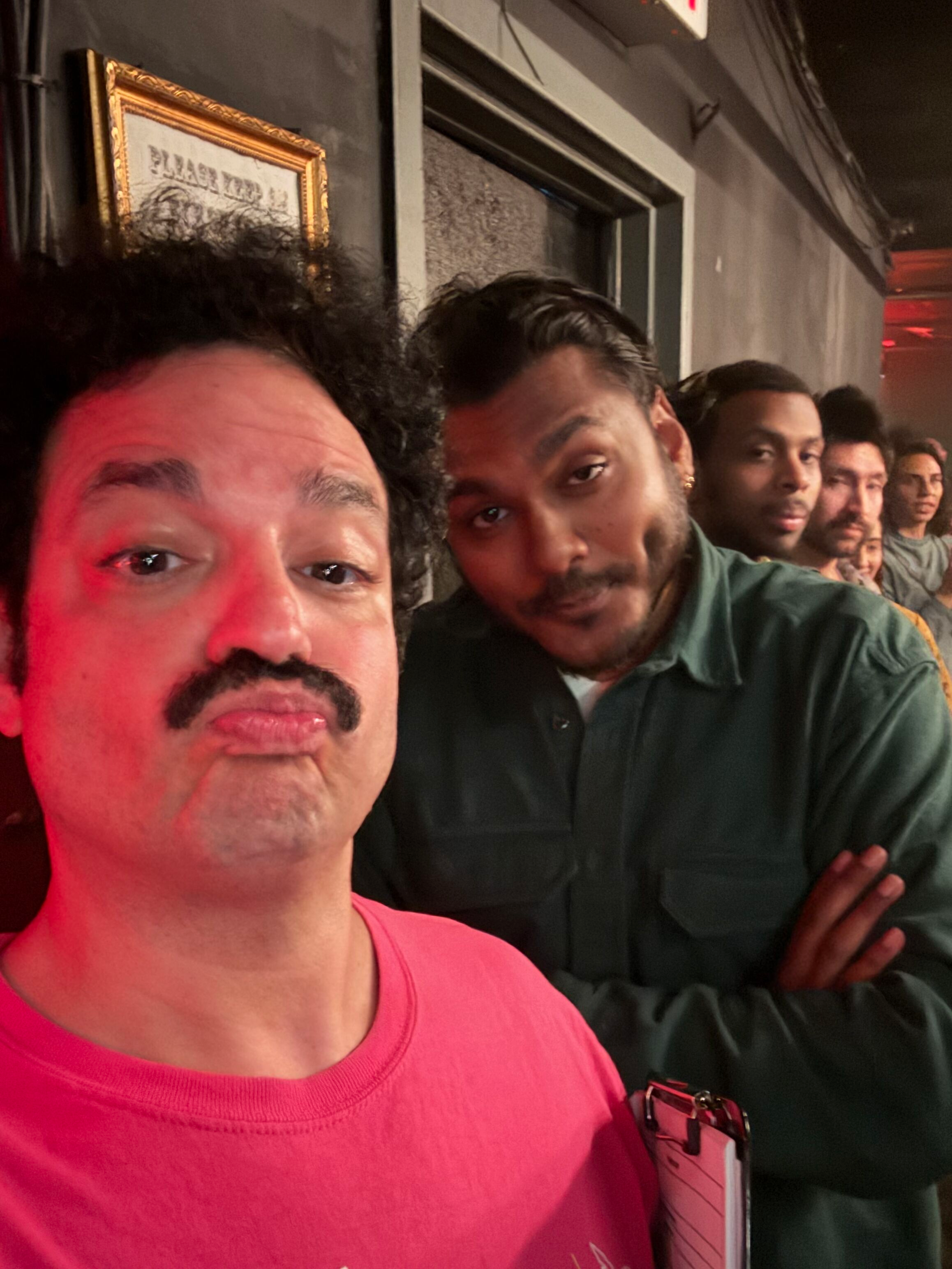 Close-up of two people taking a selfie in a dimly lit indoor setting, with a framed piece of art or poster on the wall behind them, and other people visible in the background.
