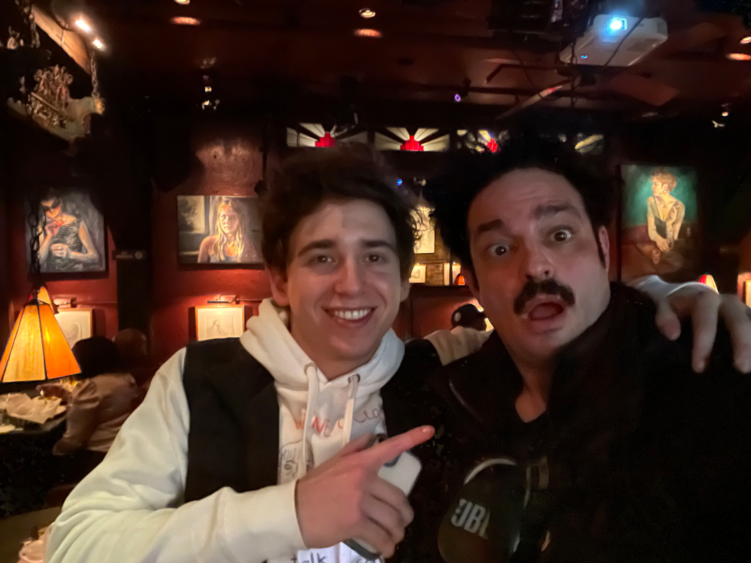 Two men taking a selfie inside a dimly lit bar or restaurant with artwork on the walls and a table lamp visible in the background.