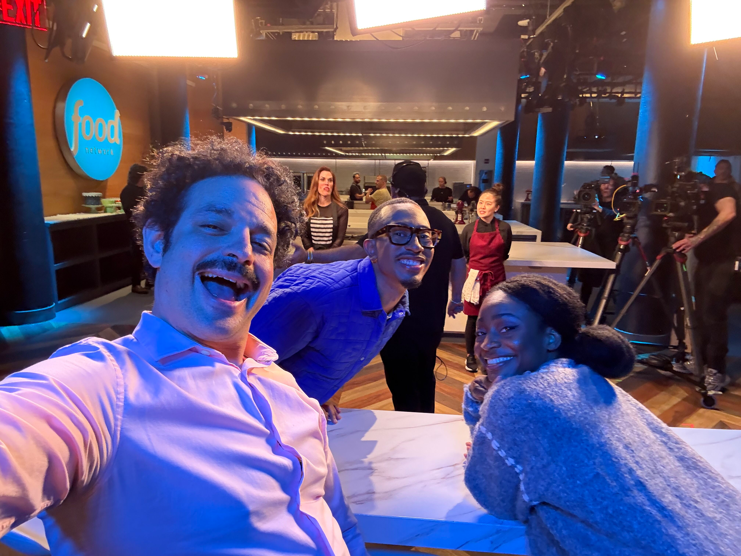 People taking a selfie at a restaurant or bar with a Food Network sign and a camera crew in the background.