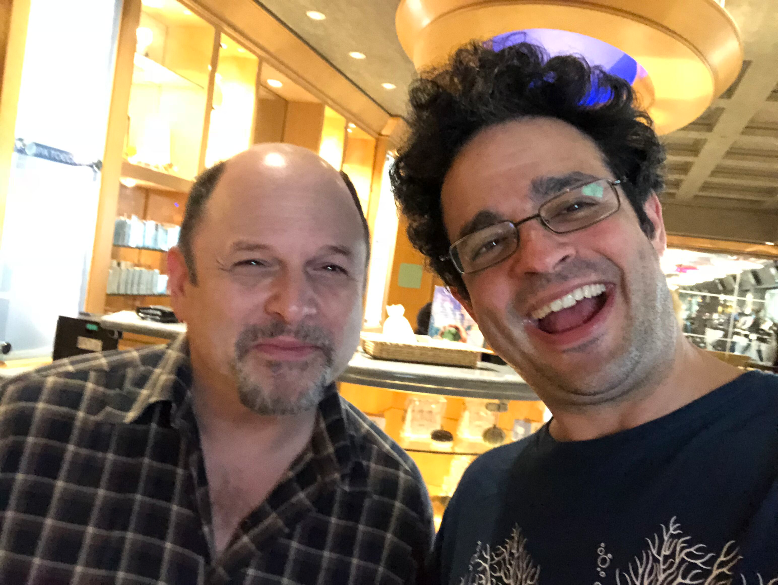 Two men are smiling and posing for a selfie in a warmly lit restaurant with yellow and brown decor. One man is wearing glasses and has curly hair, and the other has a bald head with facial hair.