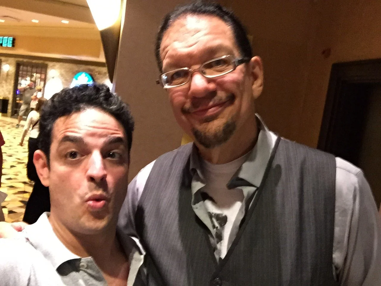 Two men taking a selfie together indoors at a social event. One man has dark hair and is making a surprised expression, while the other man has glasses and a beard, and is smiling.