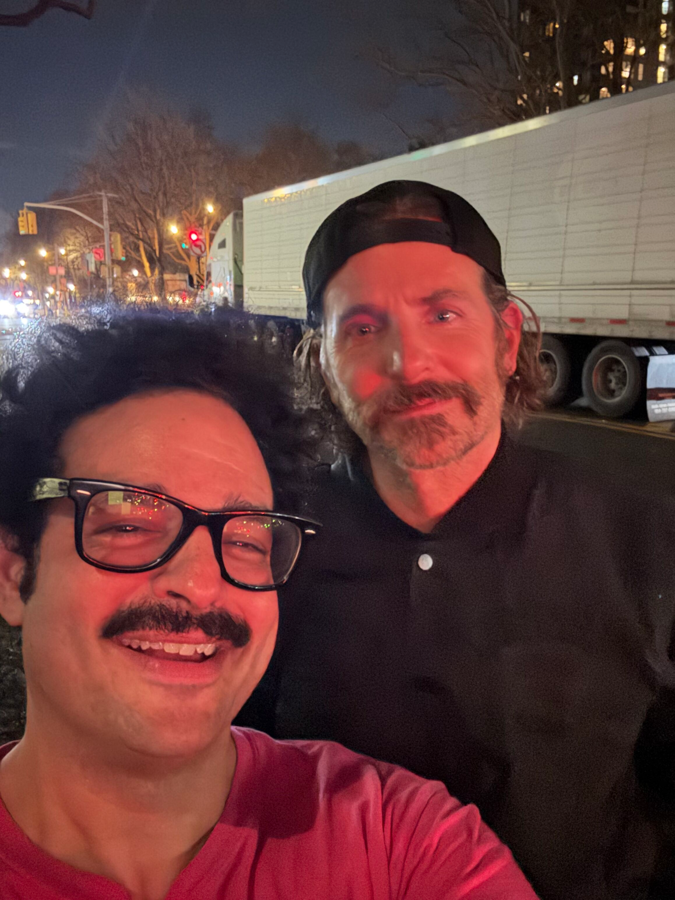 A selfie of two men smiling outdoors at night, with traffic and streetlights in the background. One man has dark hair, glasses, and a mustache, and is wearing a red shirt. The other man has light hair, a beard, and is wearing a black cap and jacket.