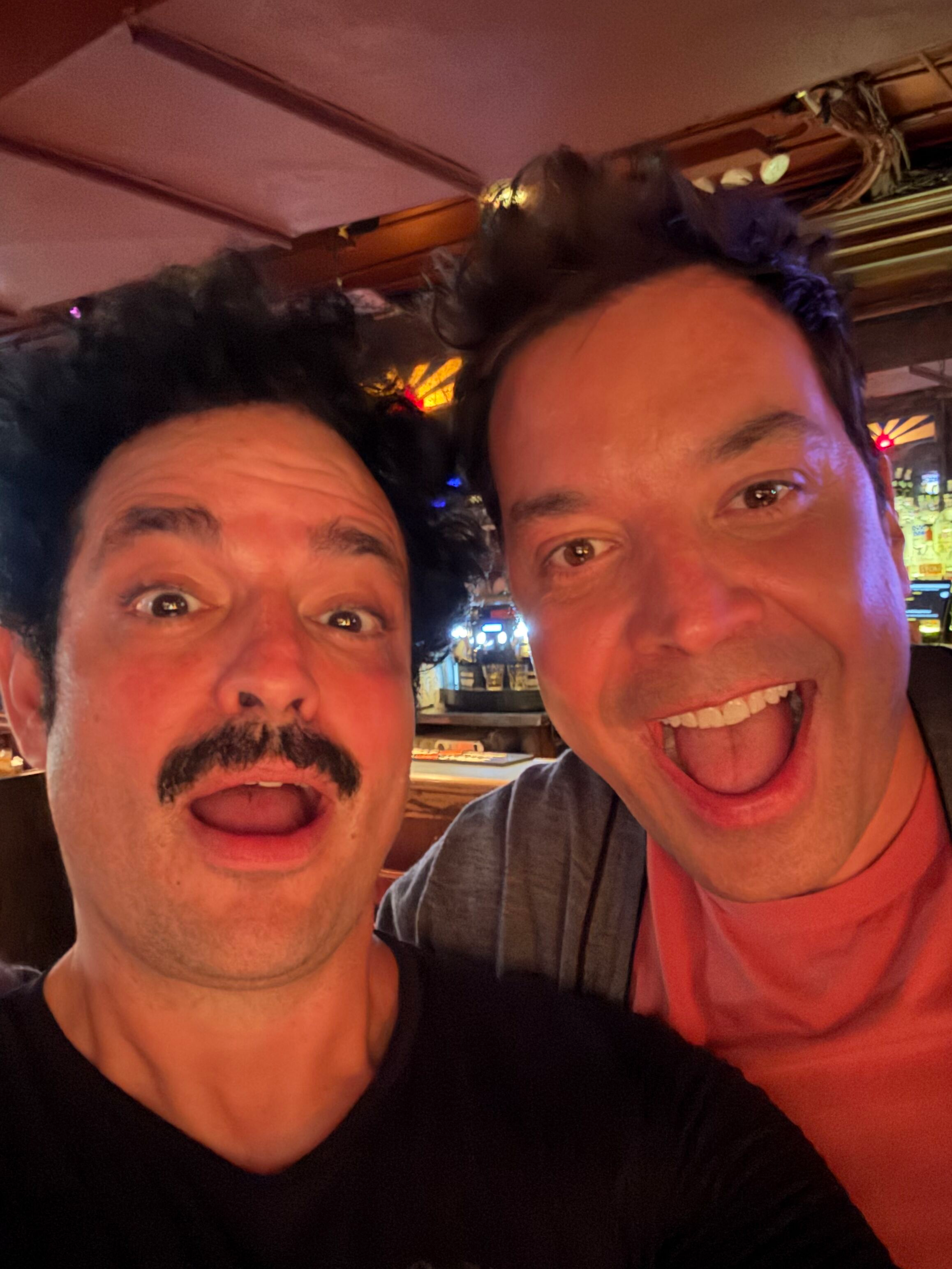 Two men are making silly faces in a dimly lit setting, possibly a bar or restaurant, with one man on the left with curly black hair and a mustache, and the other man on the right with short hair, sticking out his tongue.
