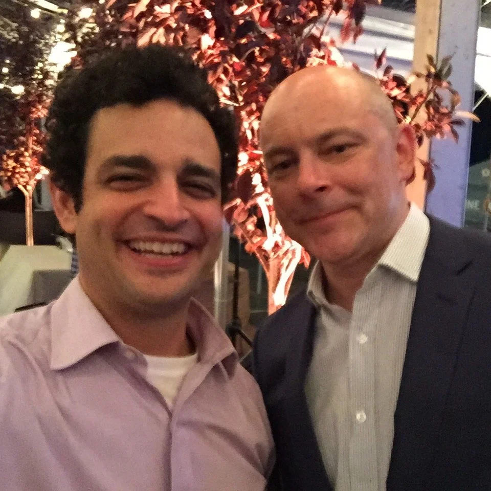 Two men smiling for a selfie outdoors at night with autumn-colored trees in the background.