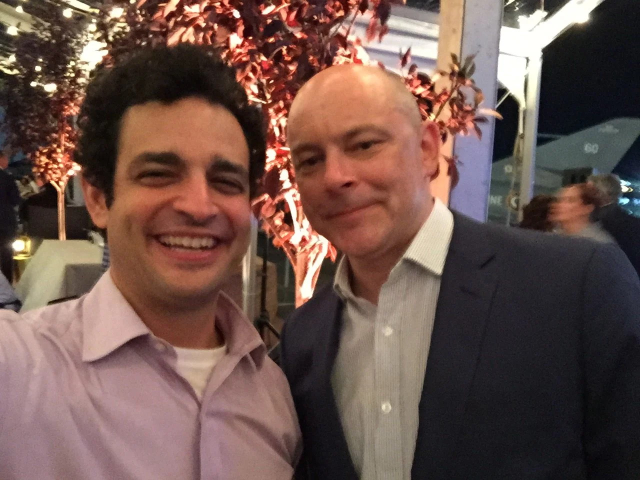 Two men smiling and posing for a photo in a dimly lit outdoor setting with a tree and string lights in the background.