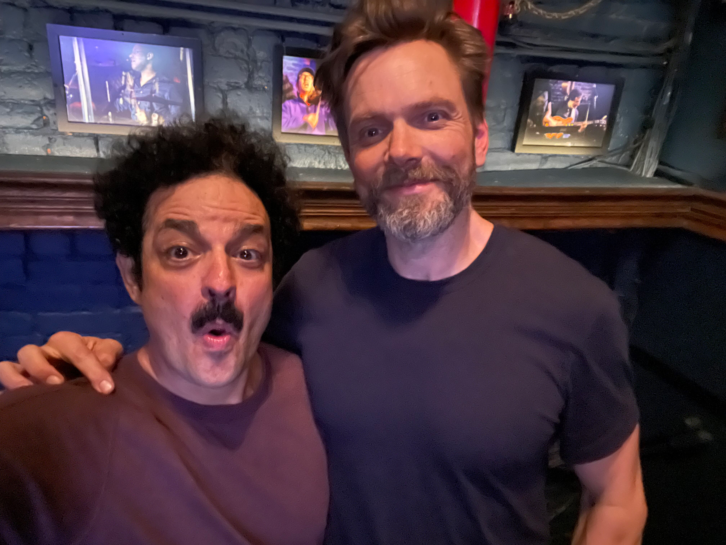 Two men taking a selfie in a dimly lit bar with framed pictures on the wall behind them.