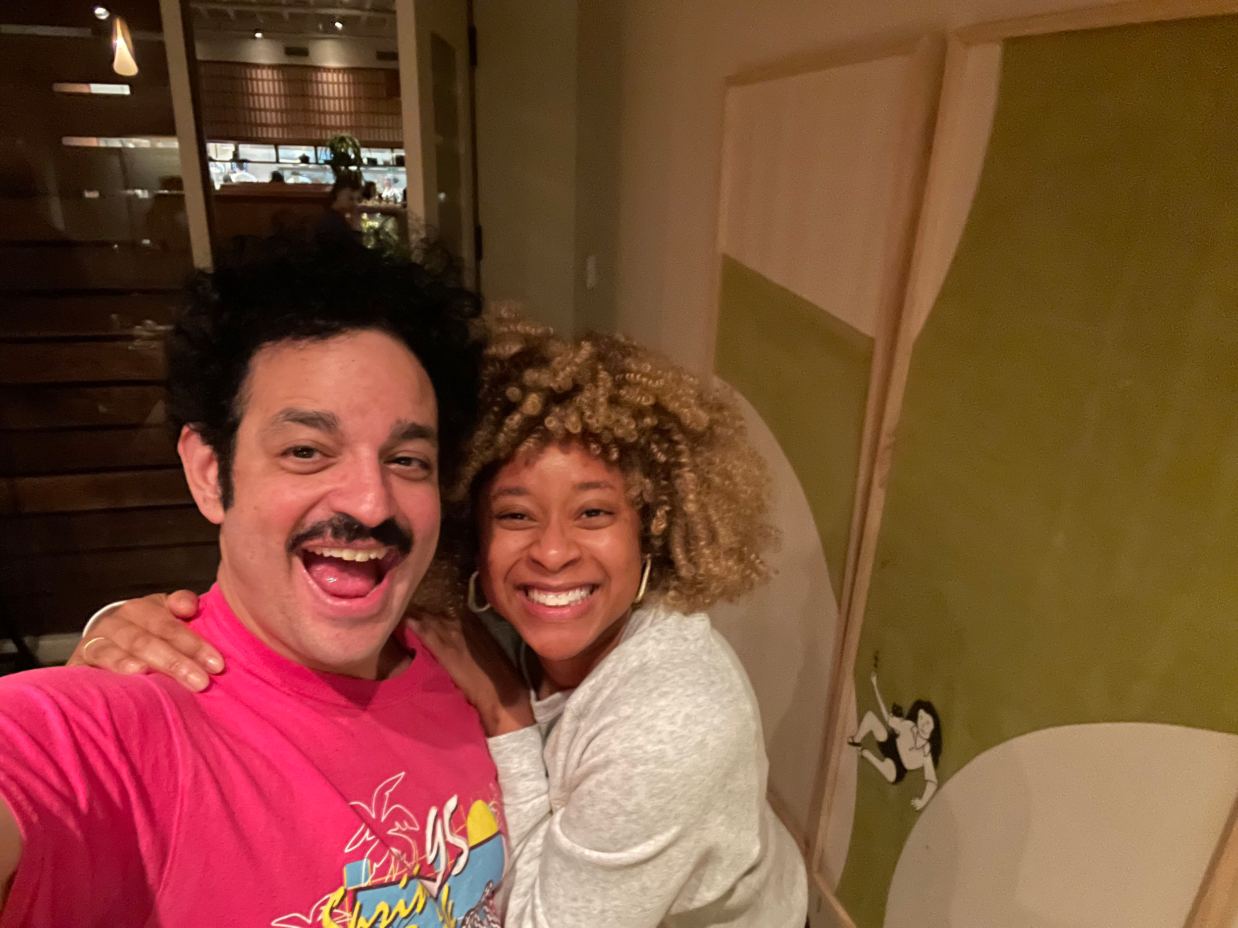 A man with black curly hair, facial hair, and wearing a bright pink shirt, and a woman with blonde curly hair and wearing a light gray shirt, are smiling and hugging each other in an indoor setting with wooden walls and a greenish ceiling.