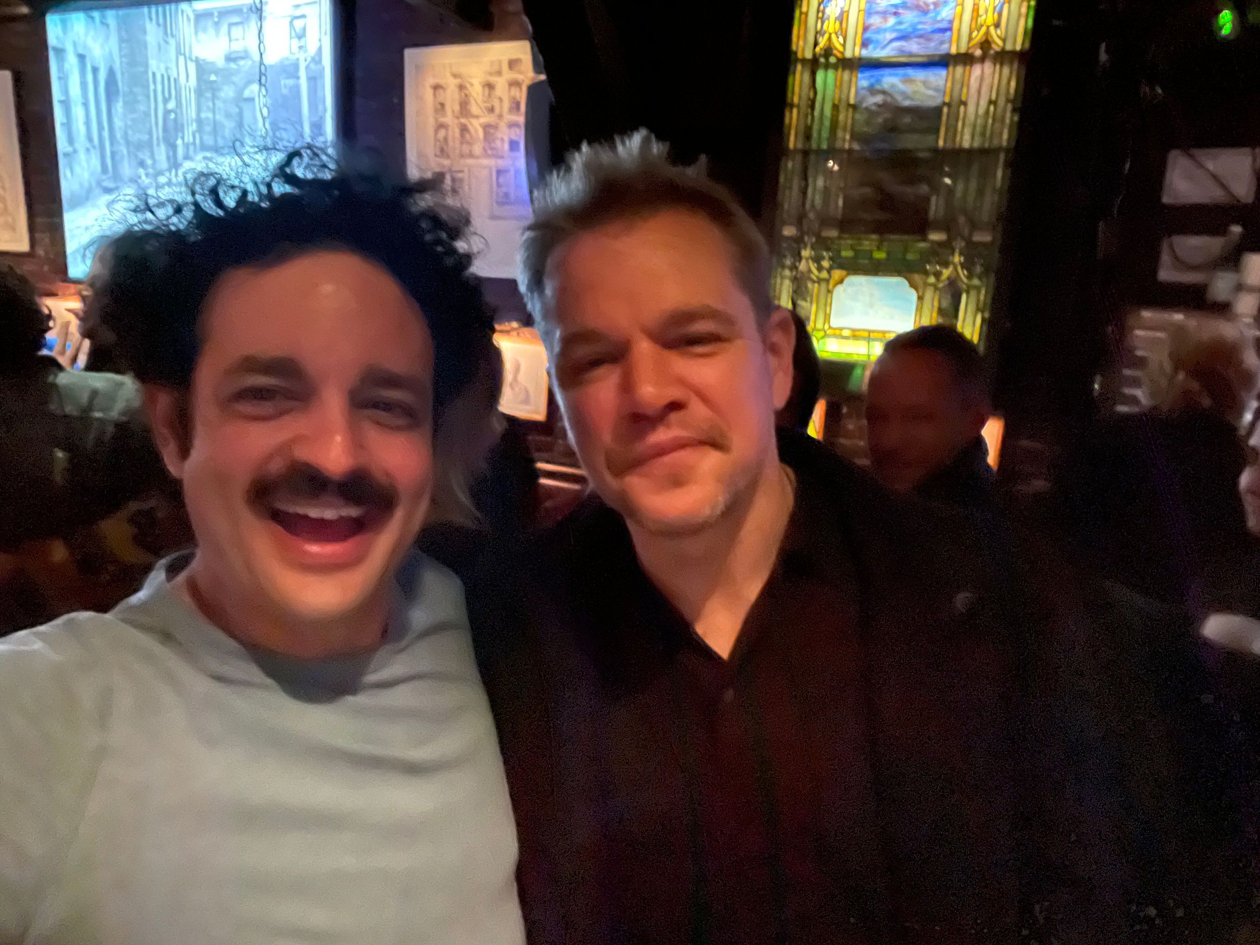 Two smiling men taking a selfie inside a dimly lit venue with stained glass windows in the background.
