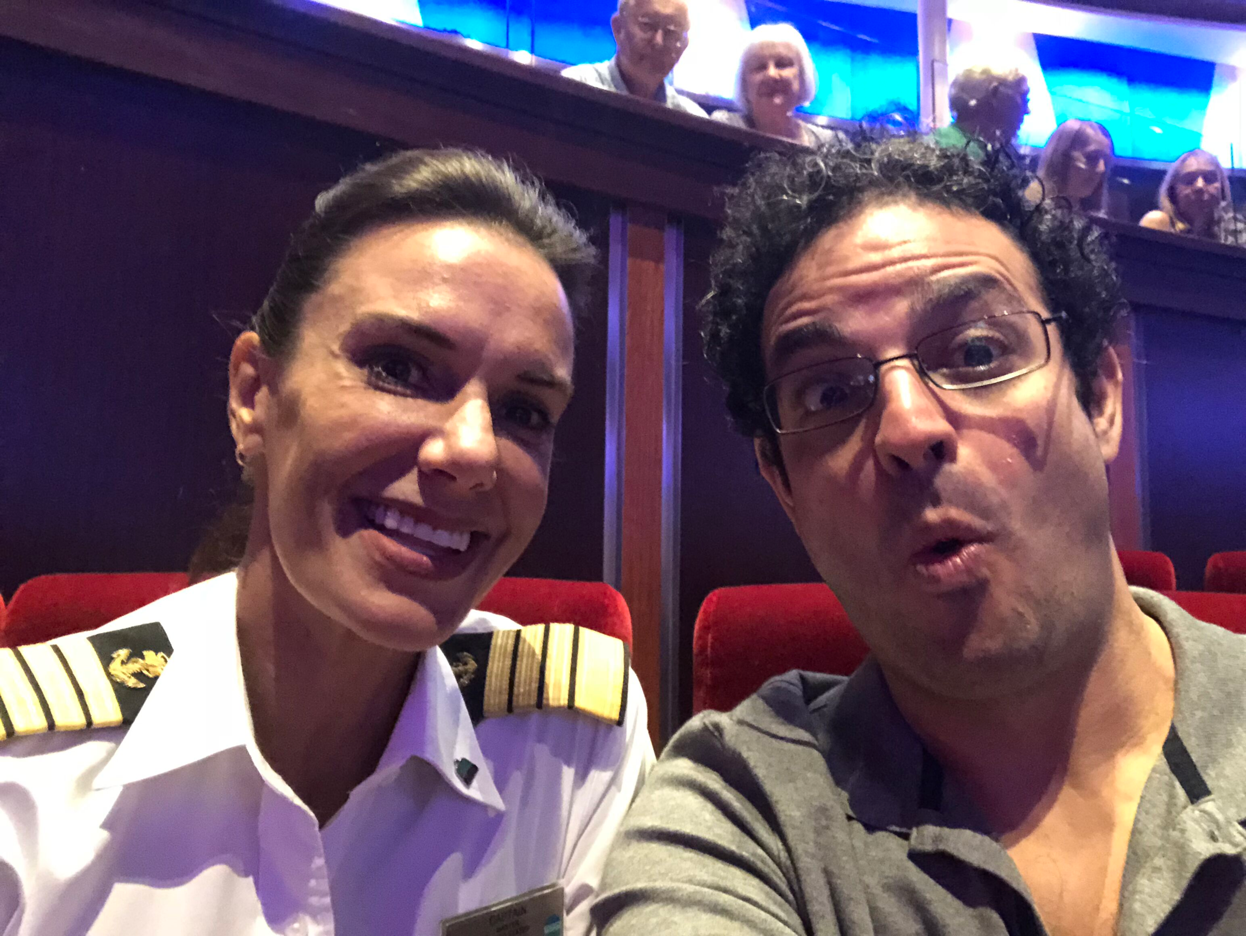 A woman with a wide smile dressed as a pilot, and a man with glasses and curly hair posing for a selfie in what appears to be an auditorium with red seats, some people are seated in the background.