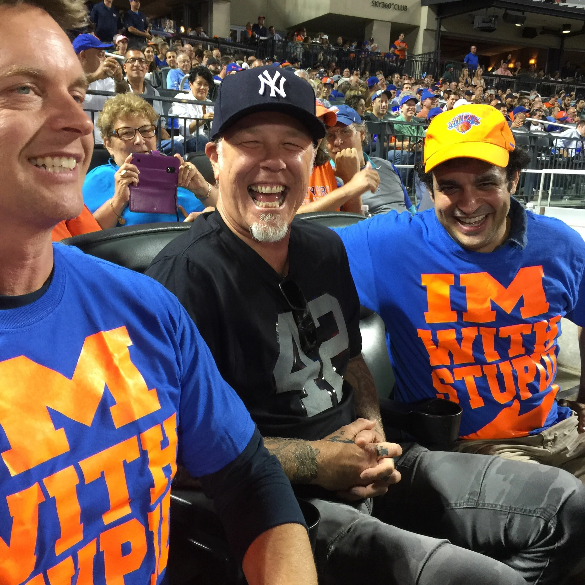 Smiling group of sports fans at a stadium, some wearing New York Yankees hats and bright orange and blue sports apparel, seated among a large crowd.