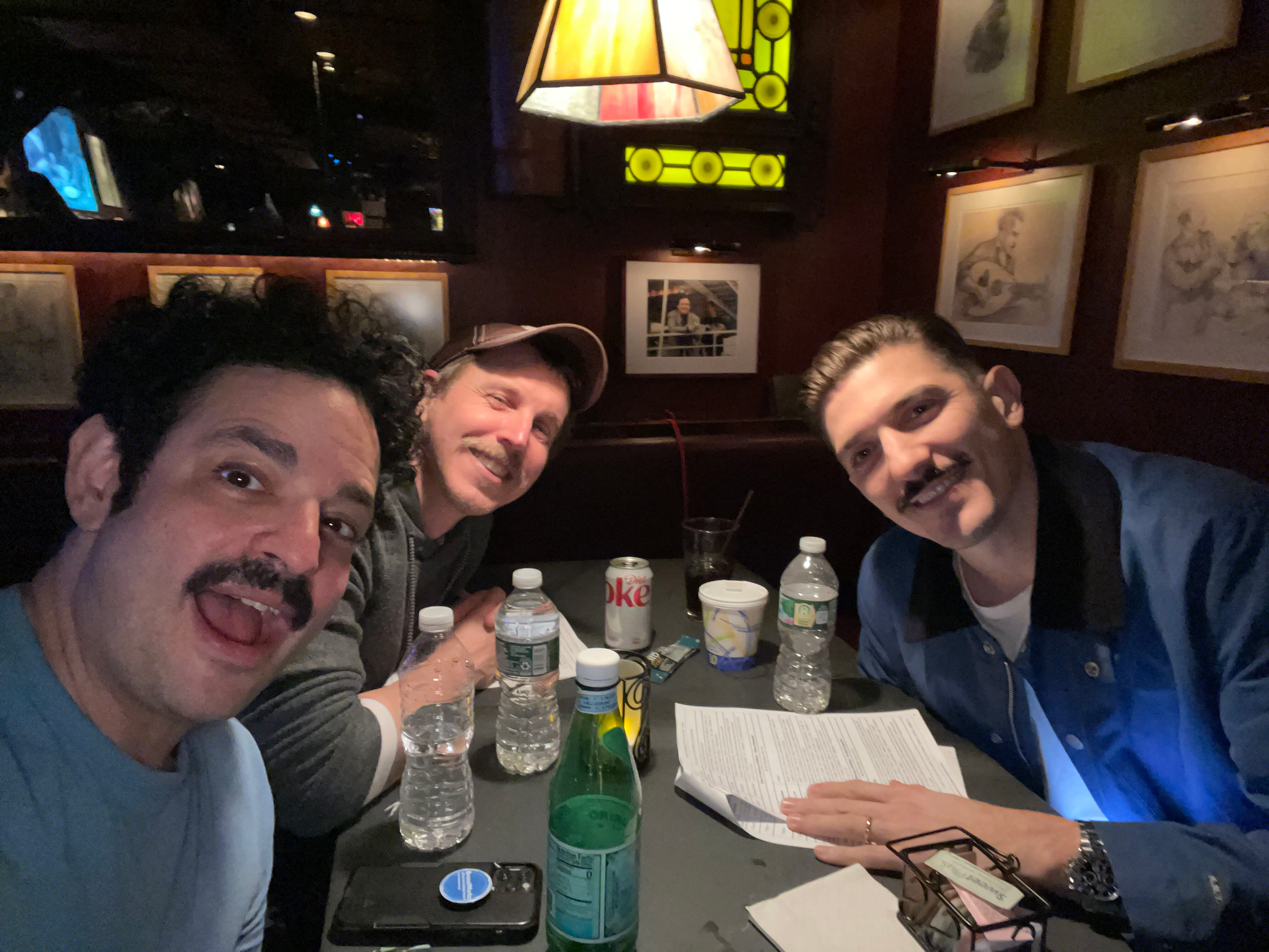 Three men sitting at a table in a dimly lit restaurant, smiling at the camera. The table has water bottles, a menu, and some drinks. Wall art and a stained glass lamp are visible in the background.
