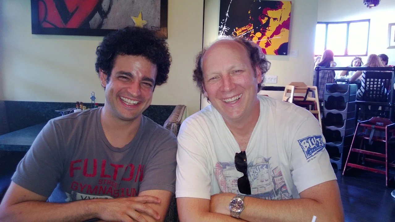 Two men sitting at a table in a restaurant, smiling at the camera. The man on the left has curly dark hair and is wearing a gray T-shirt. The man on the right has light brown hair and is wearing a white T-shirt with sunglasses hanging from the neckli