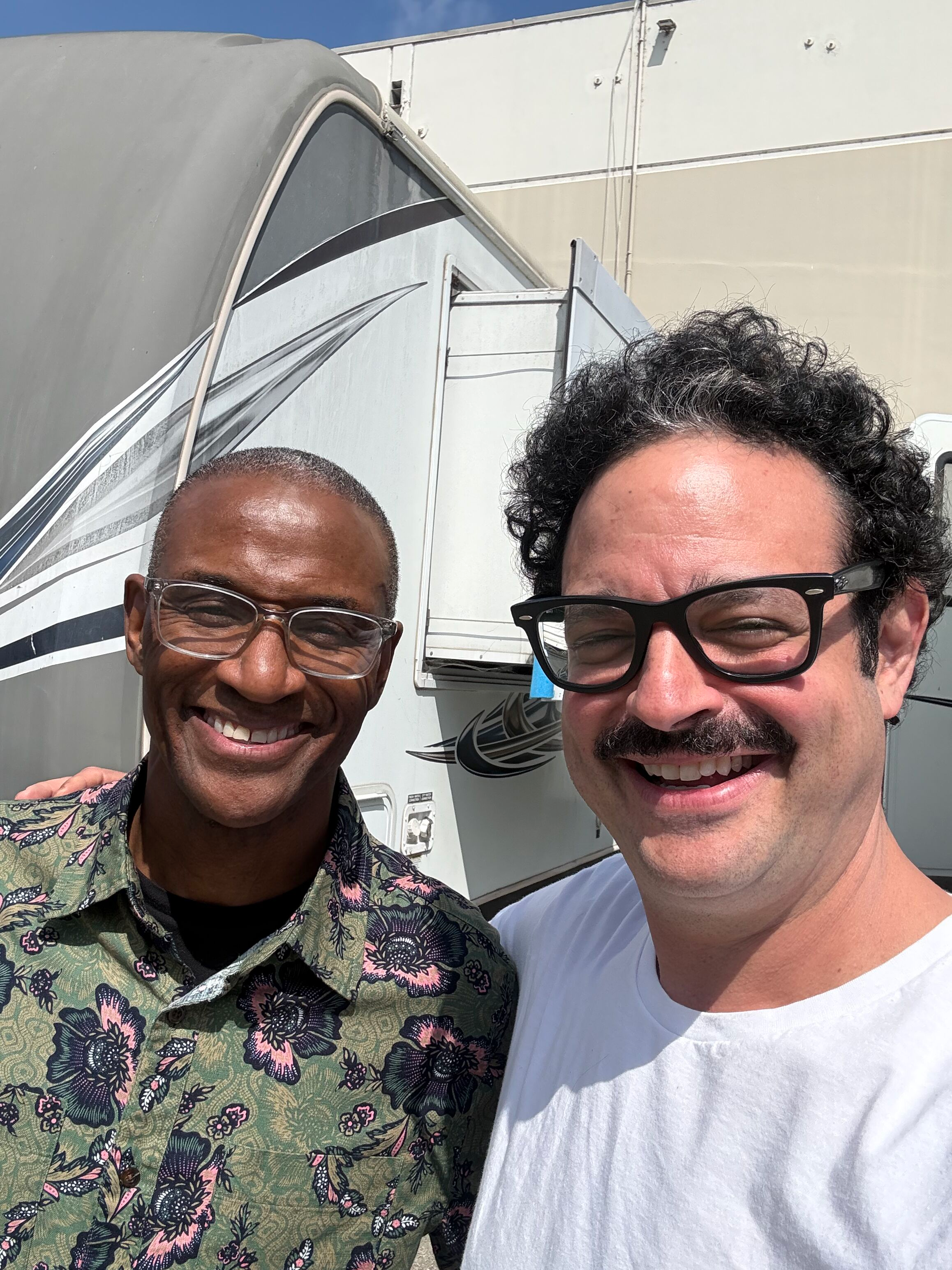 Two smiling men taking a selfie outdoors, with a large white vehicle or camper in the background. One man has dark skin, short gray hair, glasses, and a floral shirt. The other has light skin, curly black hair, glasses, and a white t-shirt.