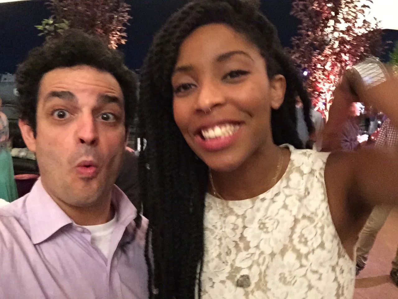 A man with dark hair and a pink shirt making an surprised face and a woman with dark curly hair wearing a white lace top smiling at the camera, in an outdoor setting at night with trees and other people in the background.