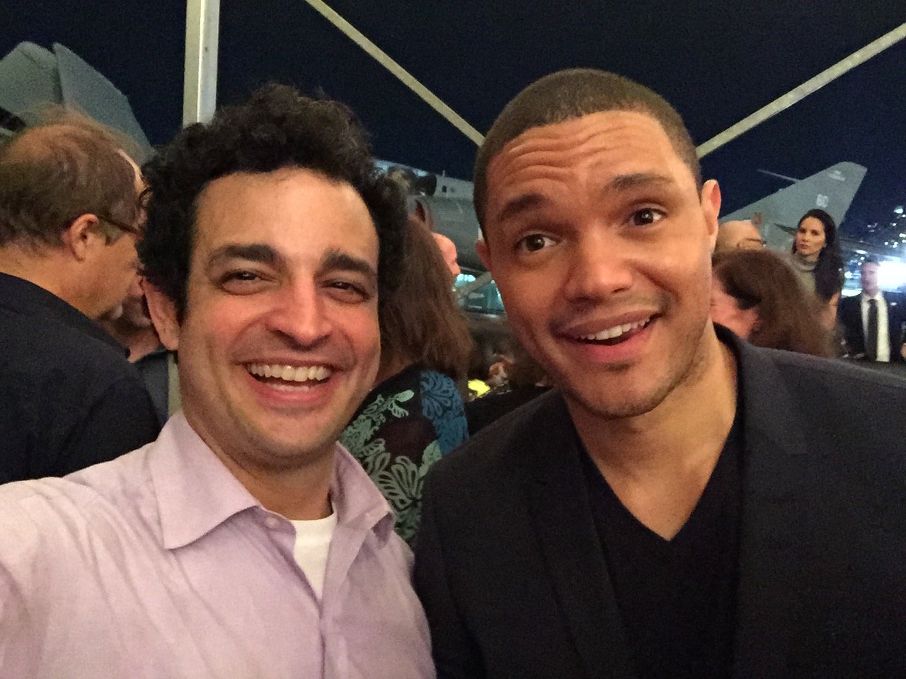 Two men smiling and taking a selfie, with airplanes and people in the background at an airport or airbase at night.