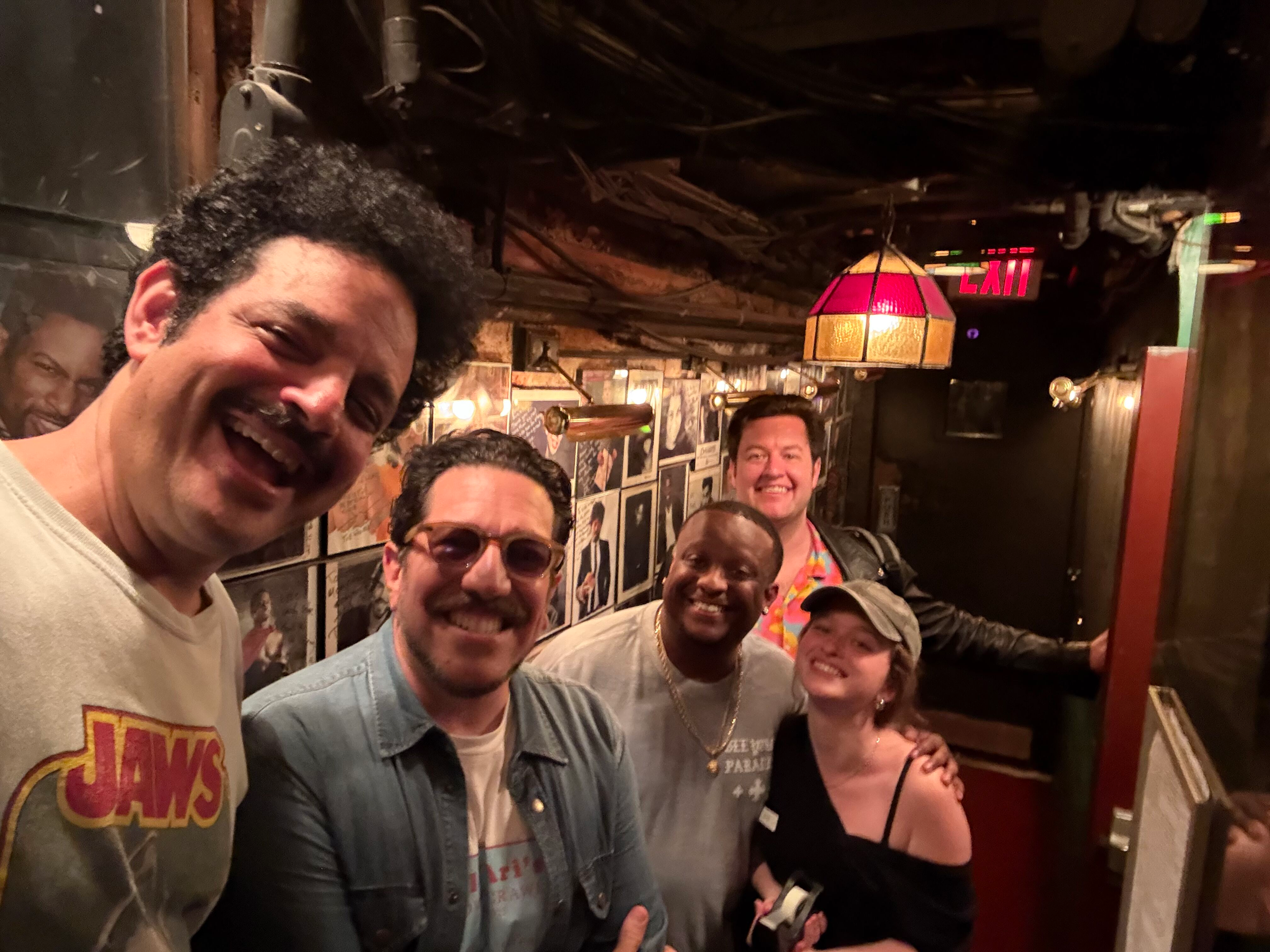 A group of people smiling and taking a selfie in a dimly lit restaurant or bar with wooden walls and framed photos. There is a red exit sign and a pink pendant lamp overhead.