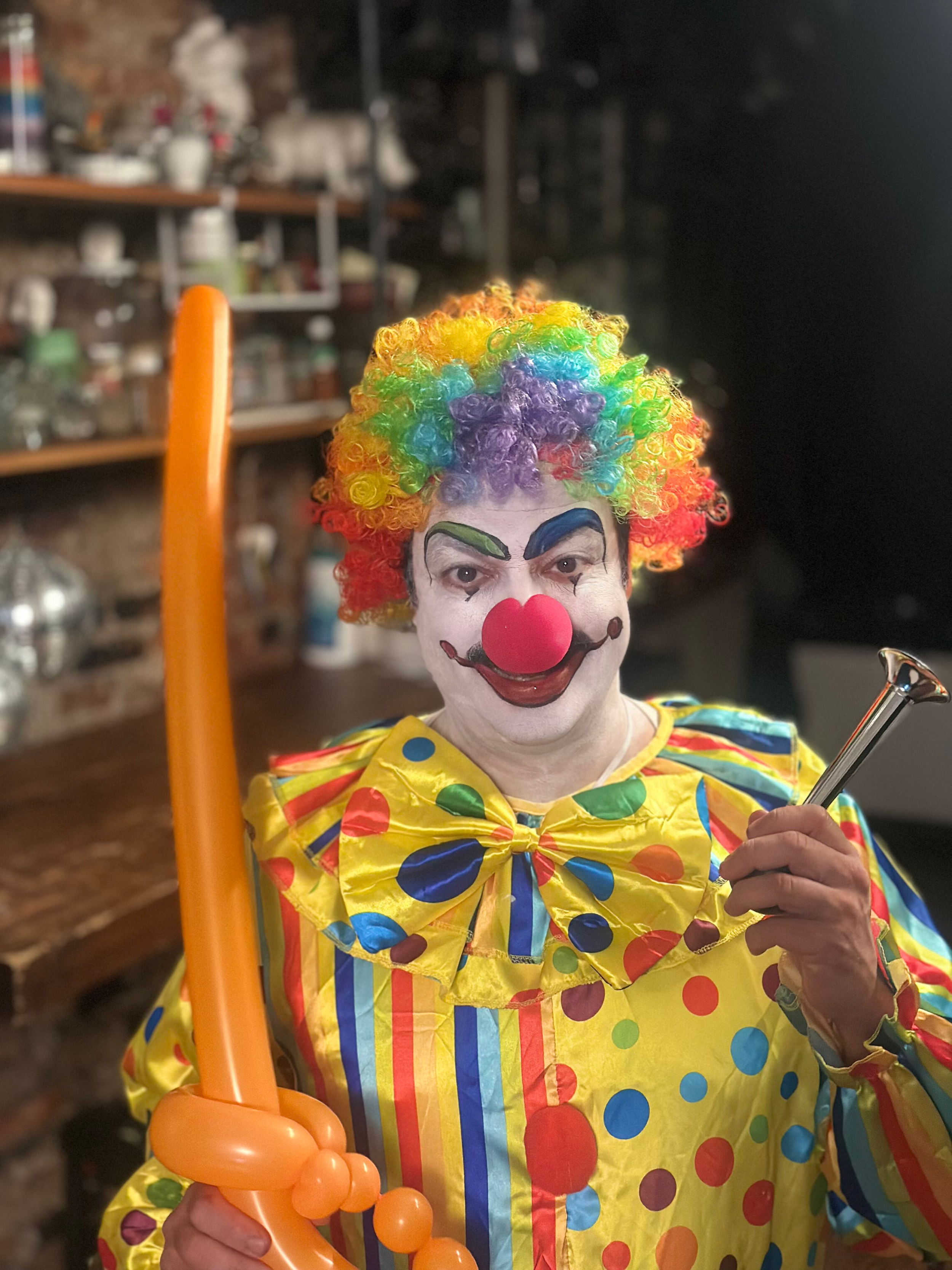 Person dressed as a clown with colorful curly wig, painted face, red nose, and a yellow outfit with multicolored polka dots, holding balloon sculpture.
