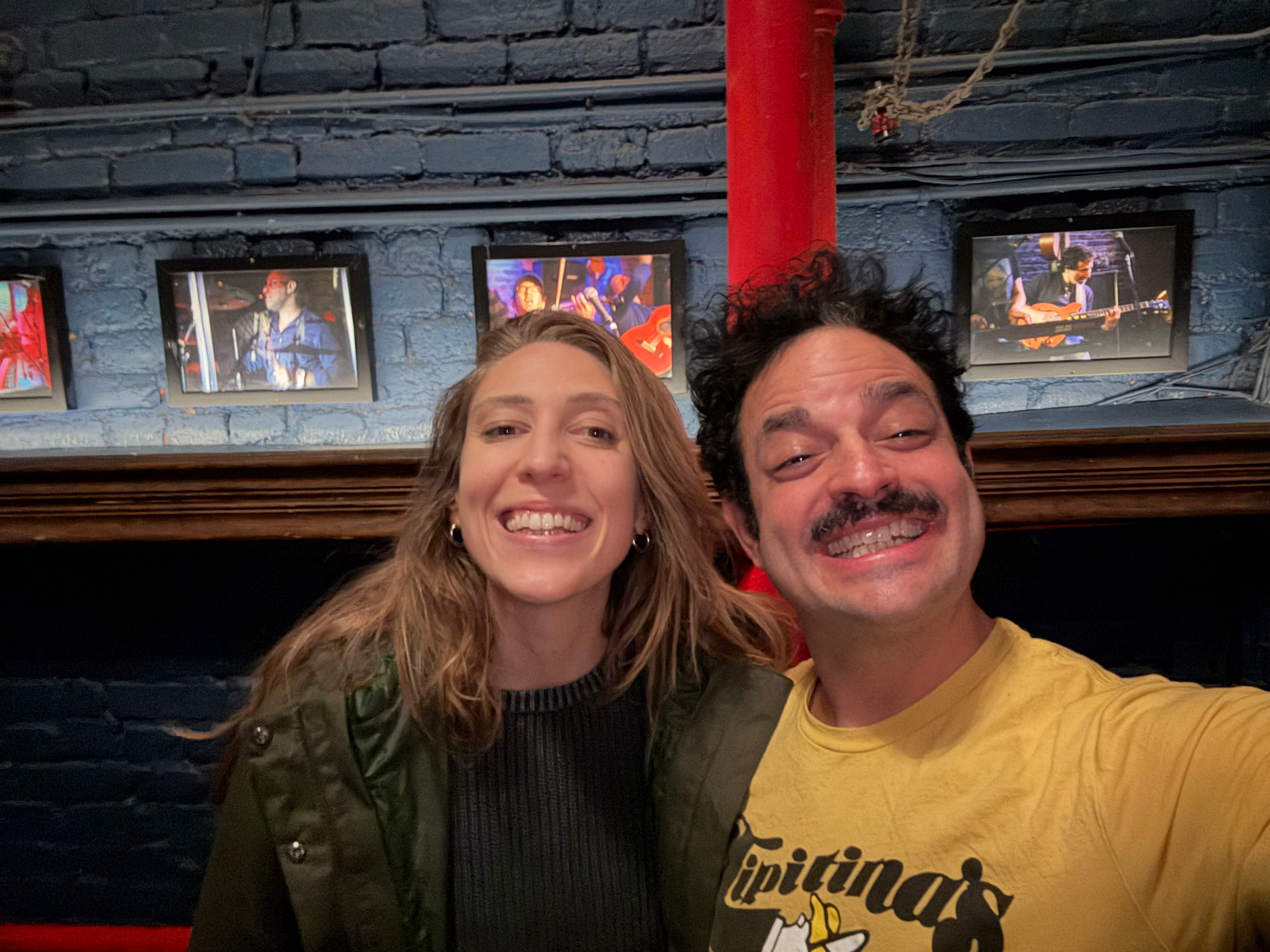 A smiling woman and man taking a selfie in front of a blue brick wall with framed photos of musicians playing instruments.